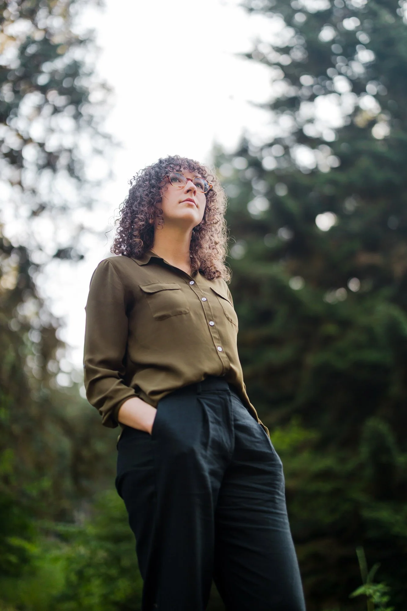 A woman with curly hair and glasses standing outdoors in a forested area, looking upward with her hands in her pockets.