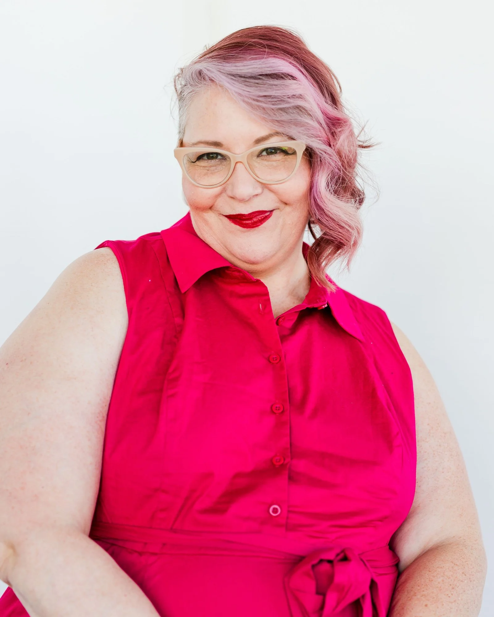 Woman with pink and purple hair wearing a sleeveless bright pink shirt, smiling, against a plain white background.