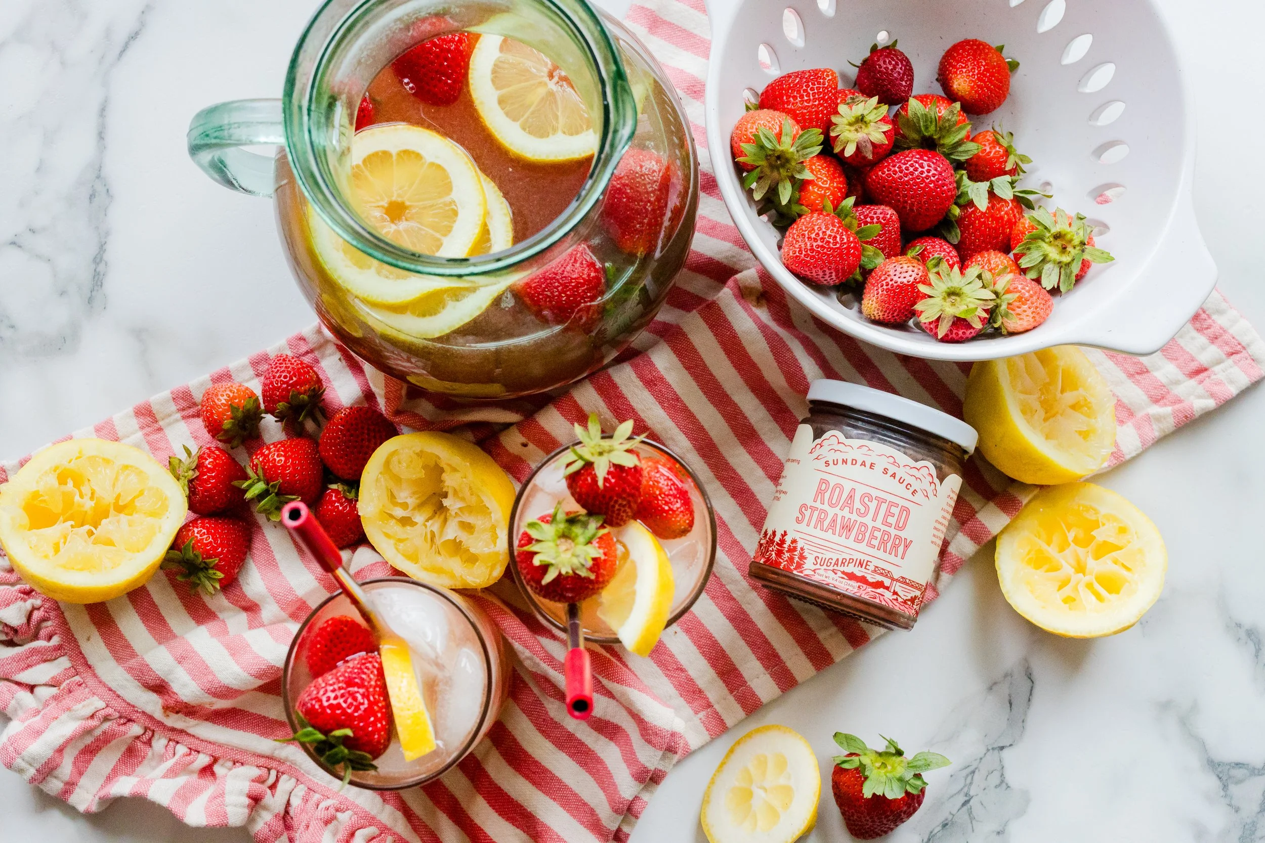 A pitcher of lemonade with lemon slices and strawberries, a bowl of fresh strawberries, halved lemons, two glasses with ice, strawberries, and lemon slices, a jar of roasted strawberry sundae sauce, all arranged on a red and white striped cloth on a 