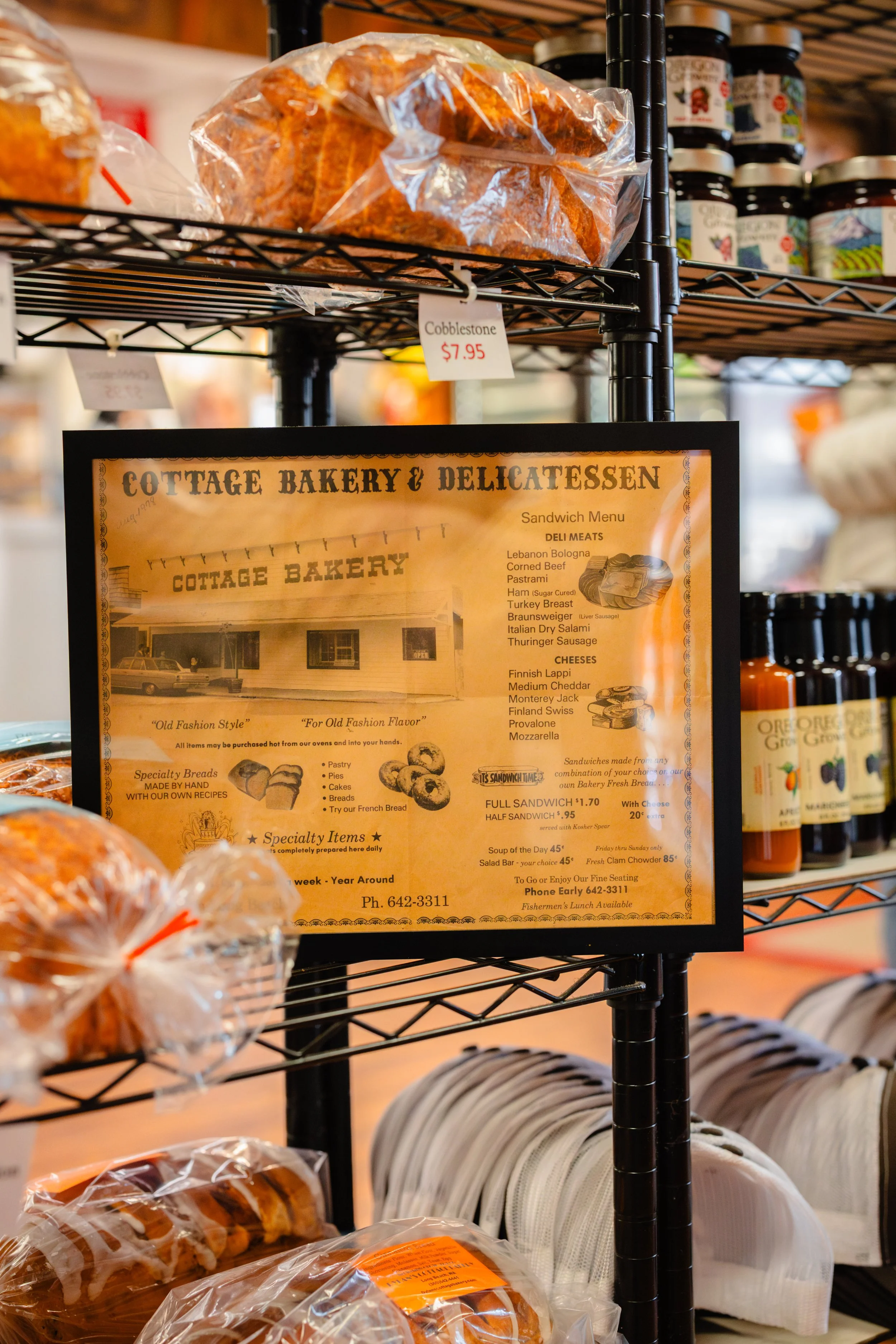 A bakery display shelf with a sign for Cottage Bakery & Delicatessen, showing prices for bread, sandwiches, and other baked goods, with packages of bread on the shelf below.