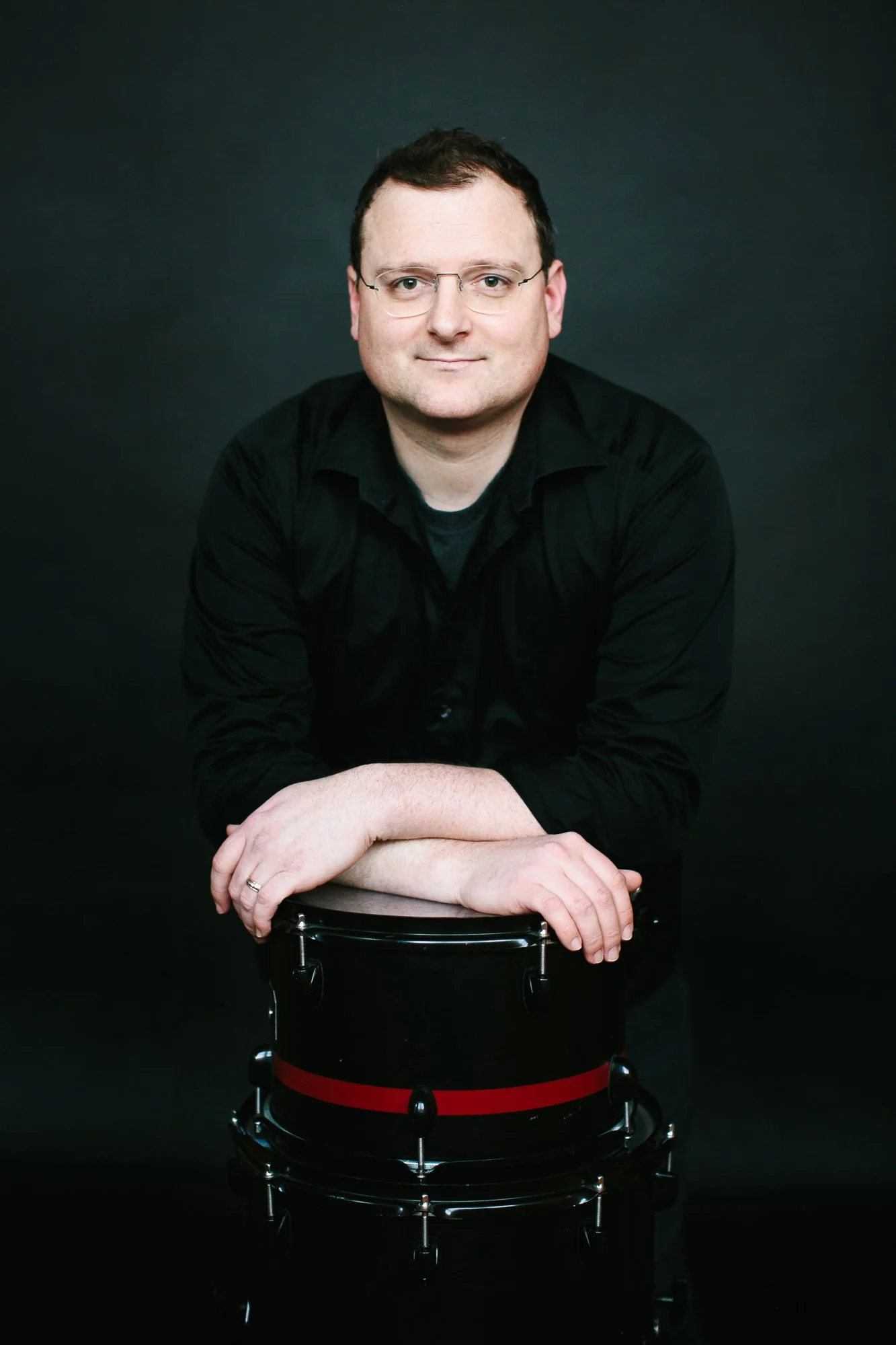 A man with glasses and short dark hair, wearing a black shirt, leaning on a black tambourine with a red stripe, against a dark background.