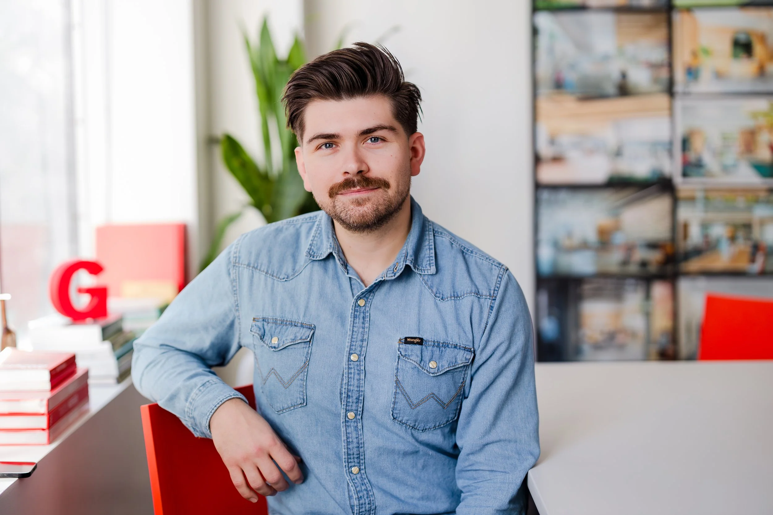 A man with dark hair and a beard sitting in a modern office or library, wearing a light denim shirt, with a slight smile, and sitting next to a white table.
