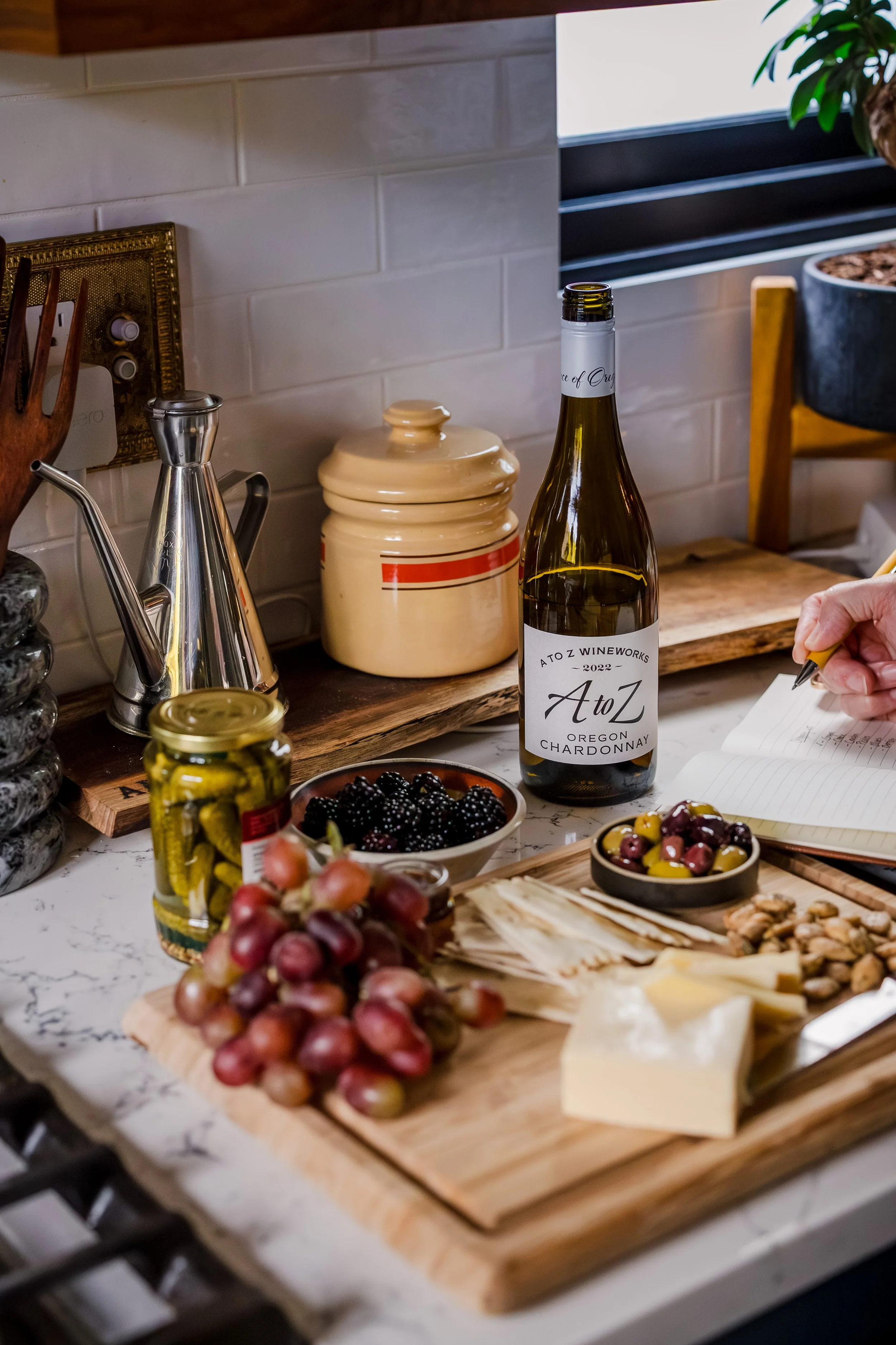 A kitchen counter with a bottle of A to Z Chardonnay wine, cheese, grapes, blackberries, olives, pickles, a jar of nuts, and a person taking notes.