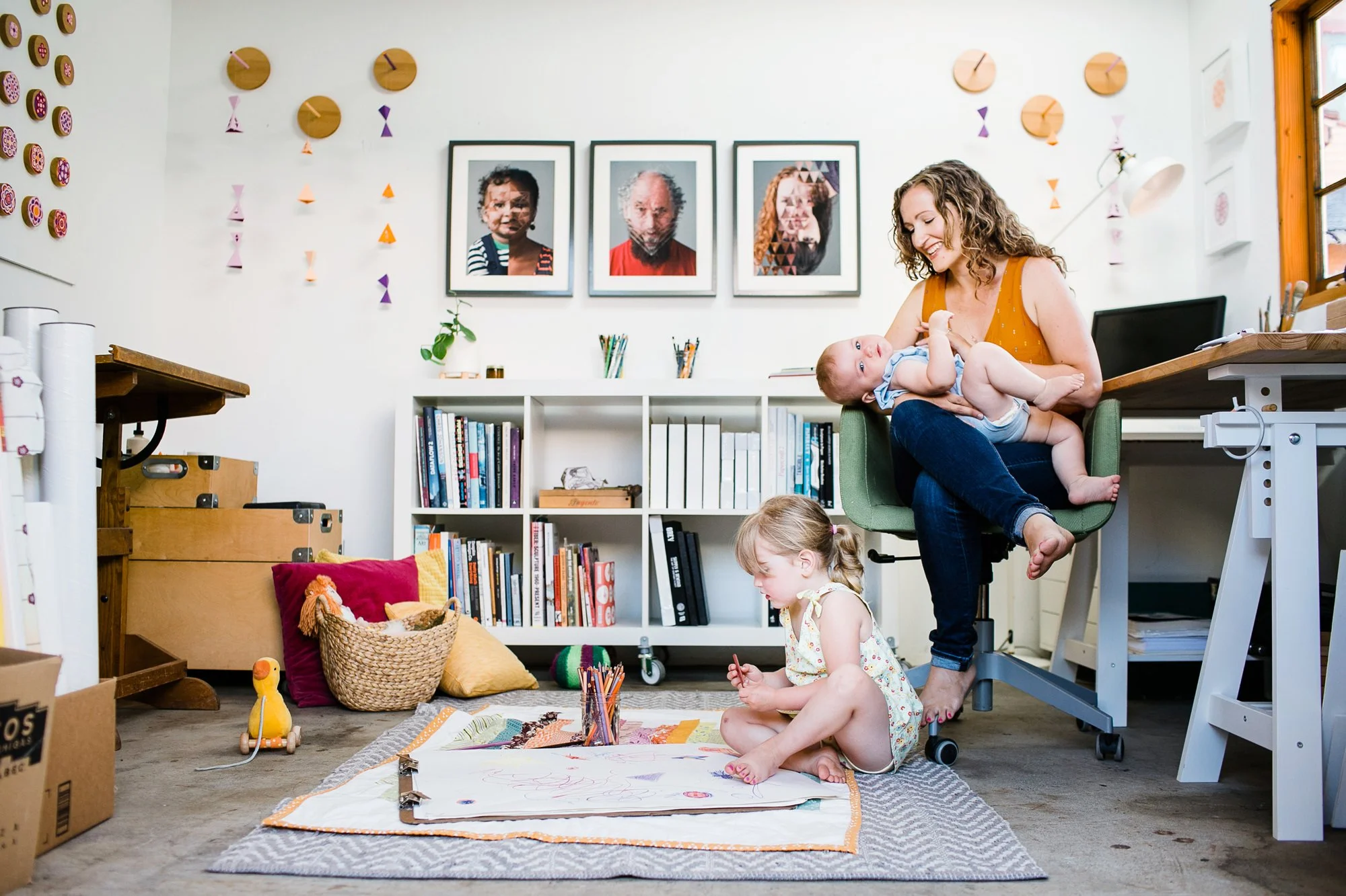 A woman sitting in a green office chair holding a baby, with a young girl sitting on the floor drawing with crayons, in a room decorated with colorful artwork and framed portraits on the wall, a bookshelf, and various toys.