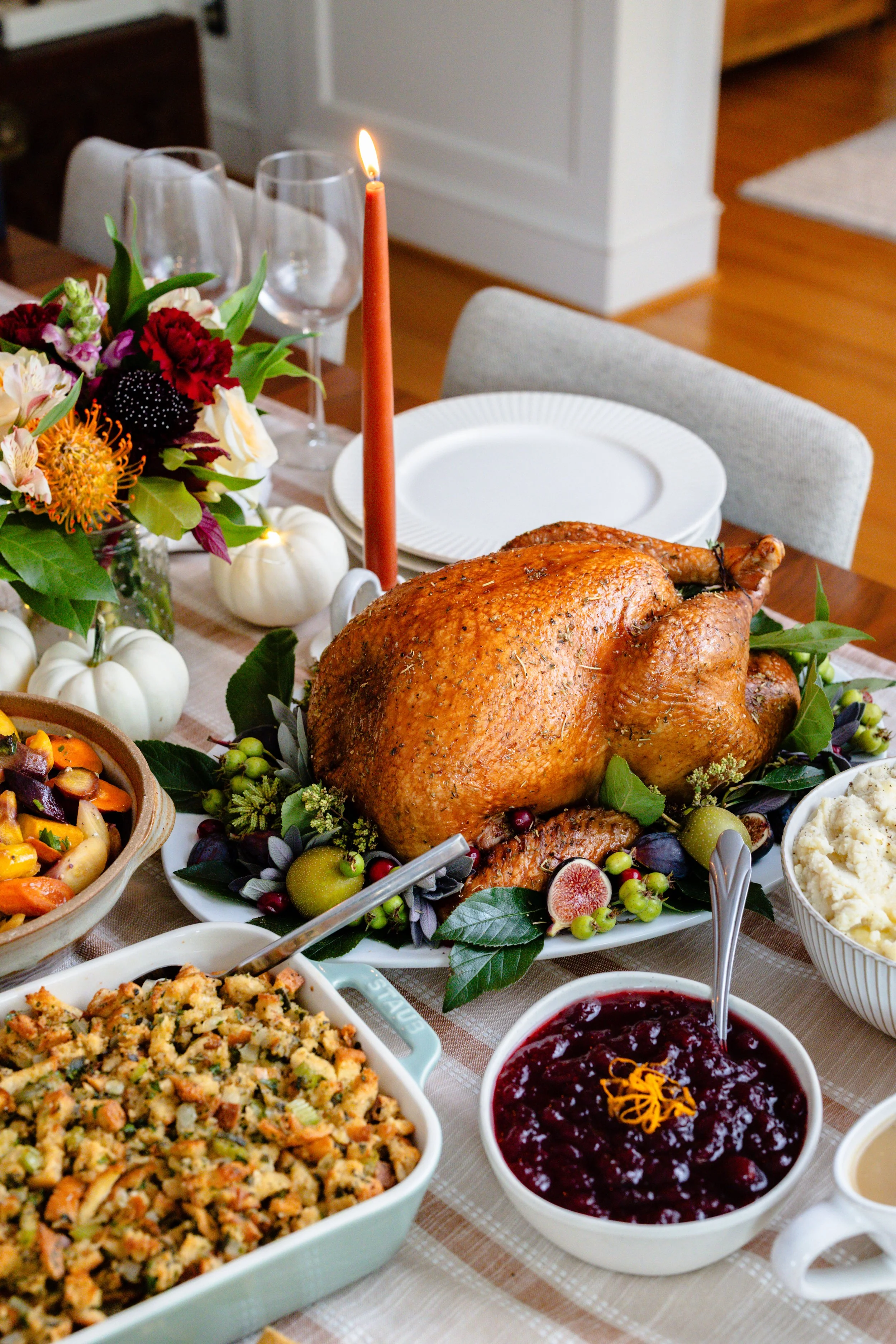 A Thanksgiving feast with a roasted turkey, stuffing, cranberry sauce, mashed potatoes, and side dishes, decorated with fall flowers and small white pumpkins.