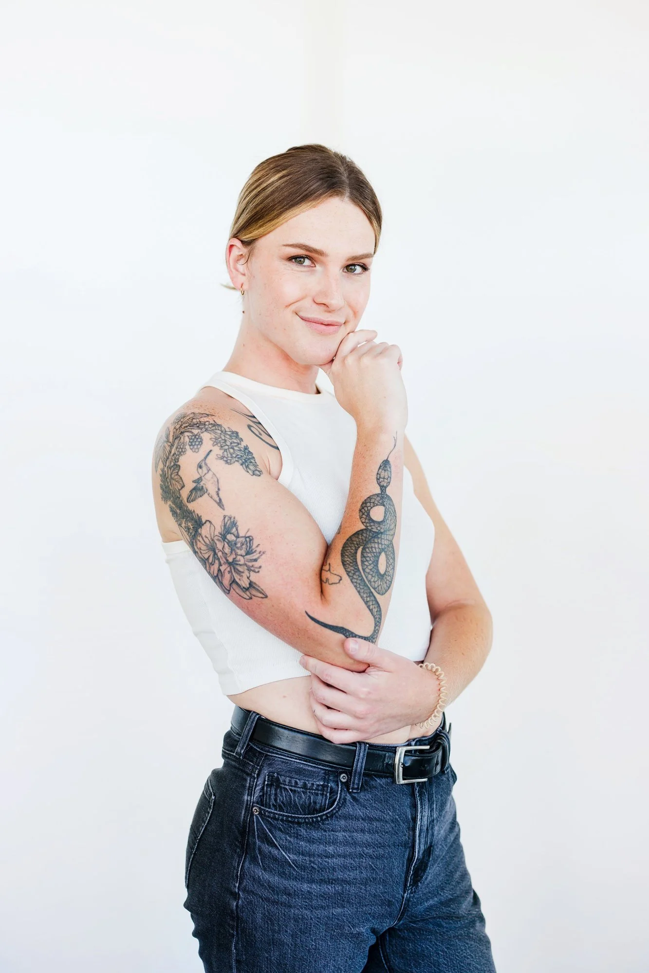 A young woman with tattoos on her arm, wearing a white sleeveless top and dark jeans, standing against a plain white background with a slight smile and her hand on her chin.