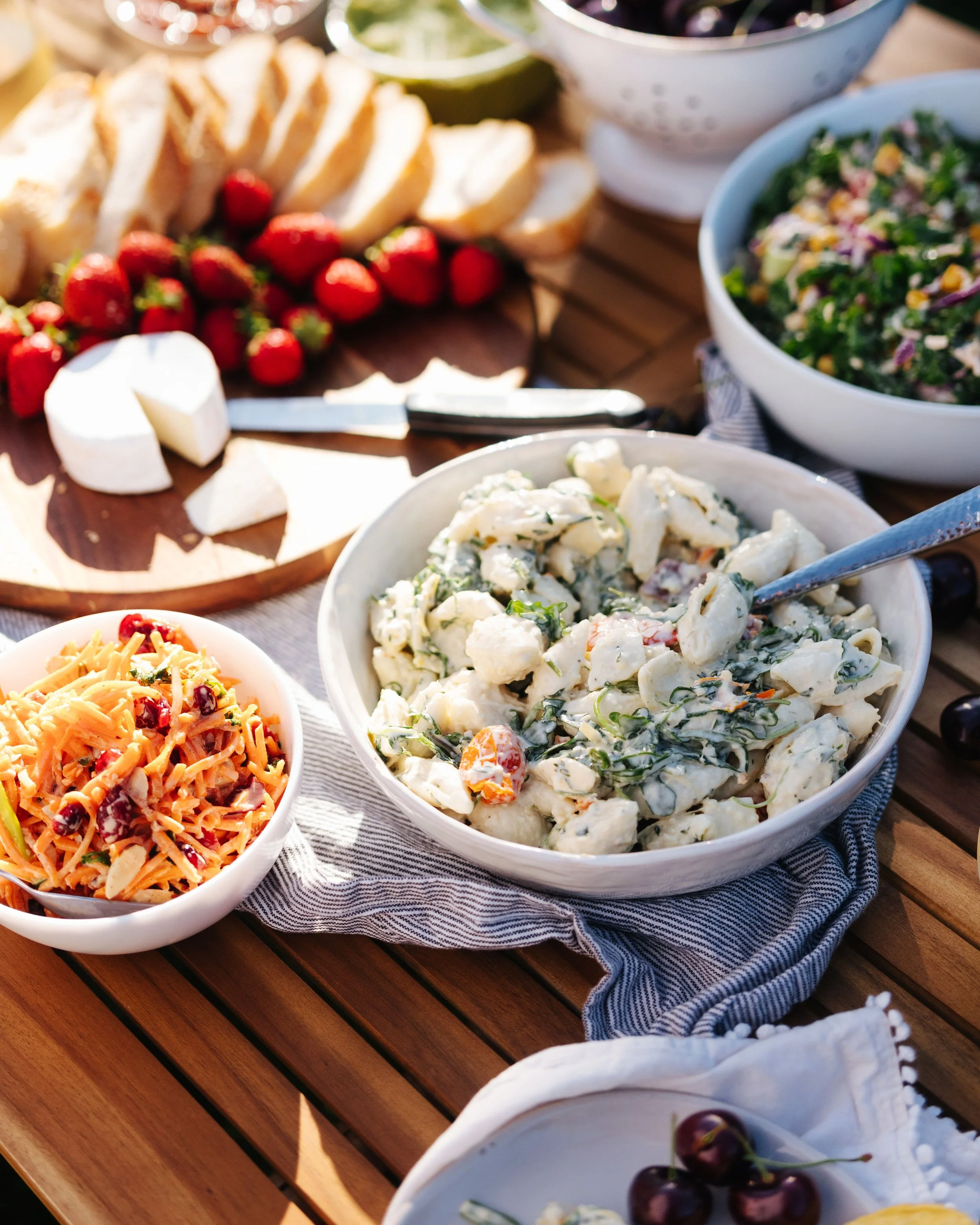 A variety of bowls and plates with salads, pasta, strawberries, cherries, bread, brie cheese, and lime slices on a wooden table for a outdoor meal.