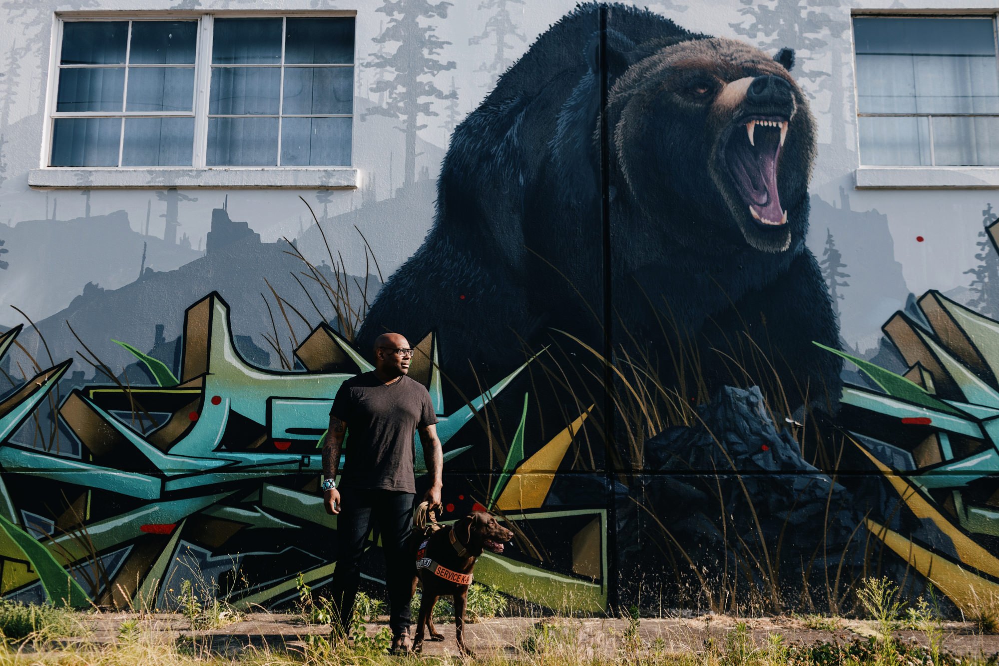 A man standing next to a service dog in front of a large colorful mural of a bear on a building wall.