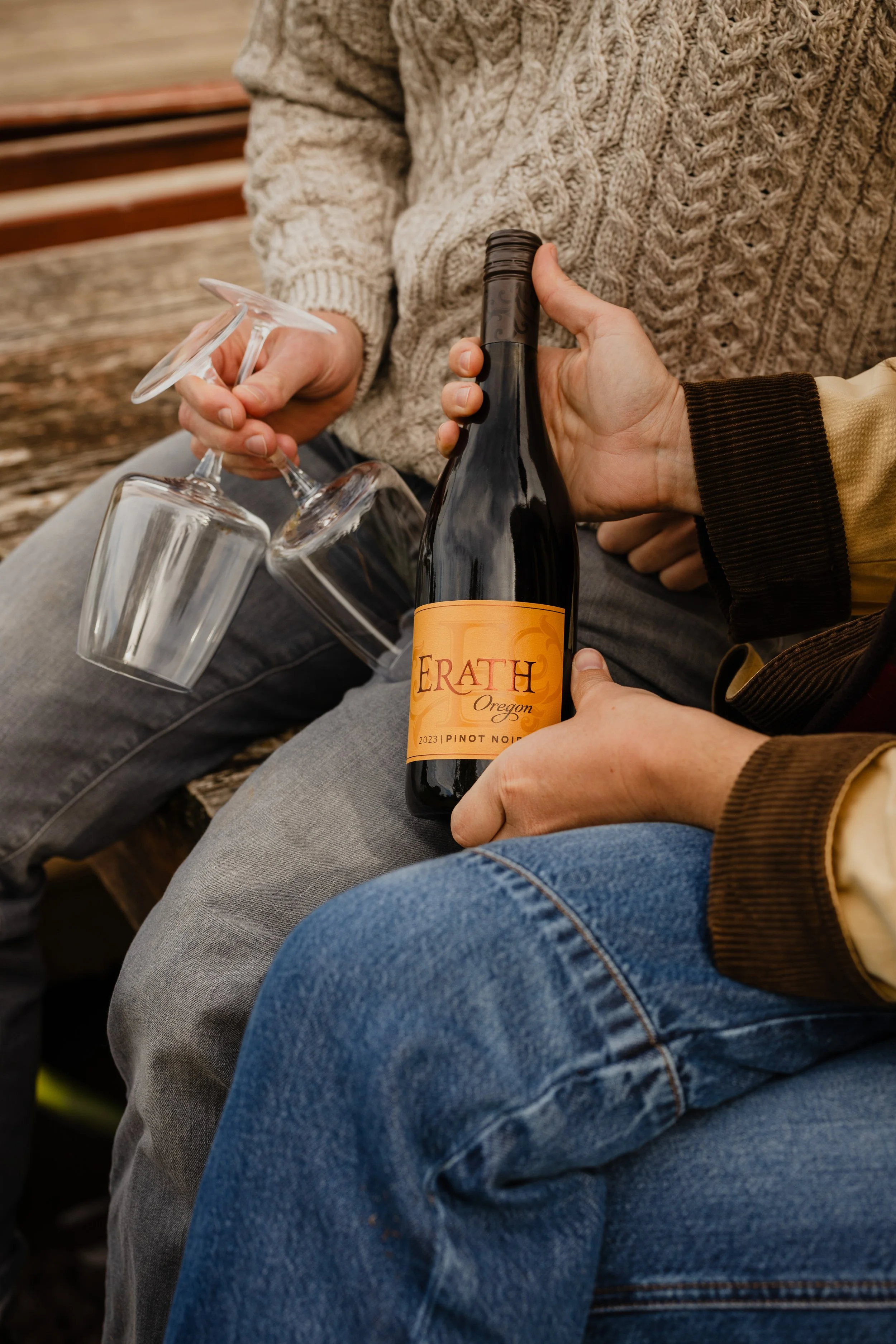Two people holding wine glasses and a bottle of Erath Oregon Pinot Noir wine, sitting on a wooden bench outdoors.