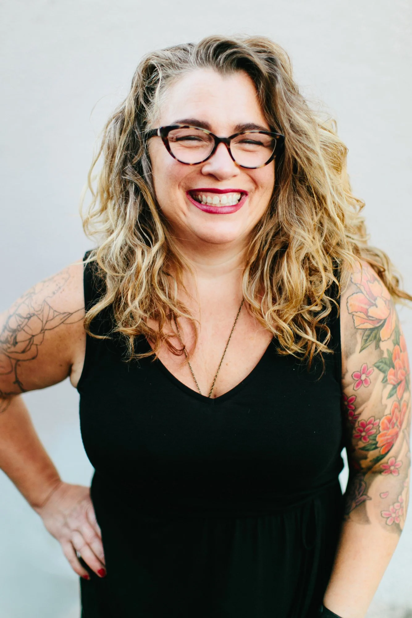 A smiling woman with curly blonde hair, wearing glasses and a black sleeveless dress, standing with her hand on her hip against a plain background.