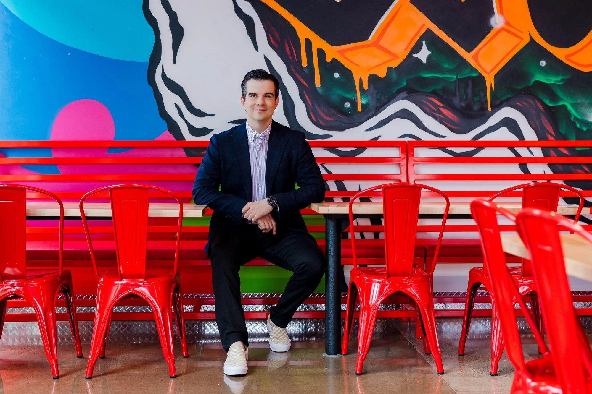 A man sitting at a table in a colorful, modern restaurant with a vibrant mural on the wall behind him. The mural features abstract shapes and bright colors, with red chairs and wooden tables surrounding him.