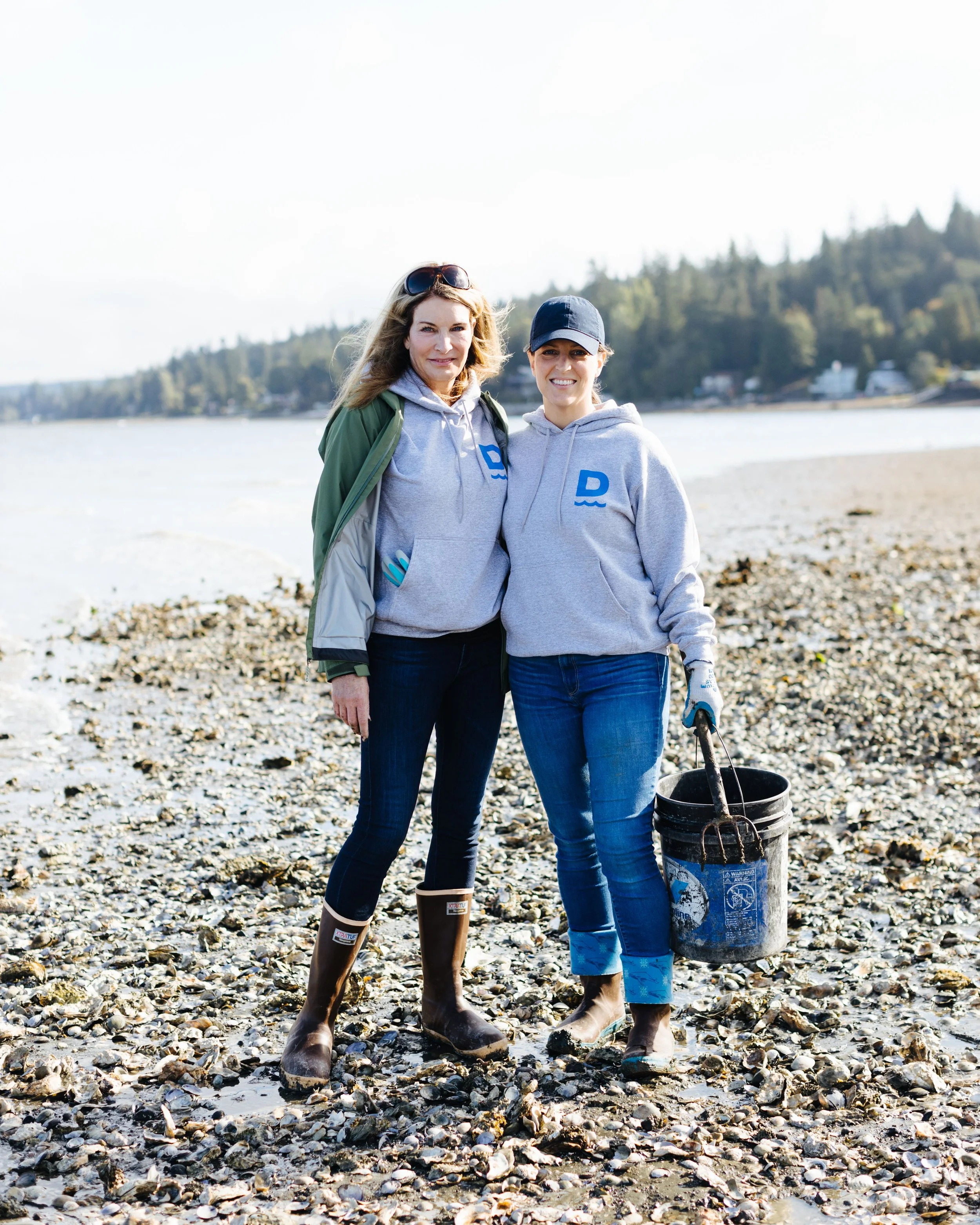 Two women standing on a rocky beach, smiling at the camera, dressed in grey hoodies, jeans, and rain boots. One woman is holding a bucket with a rake, and there are trees and houses in the background.