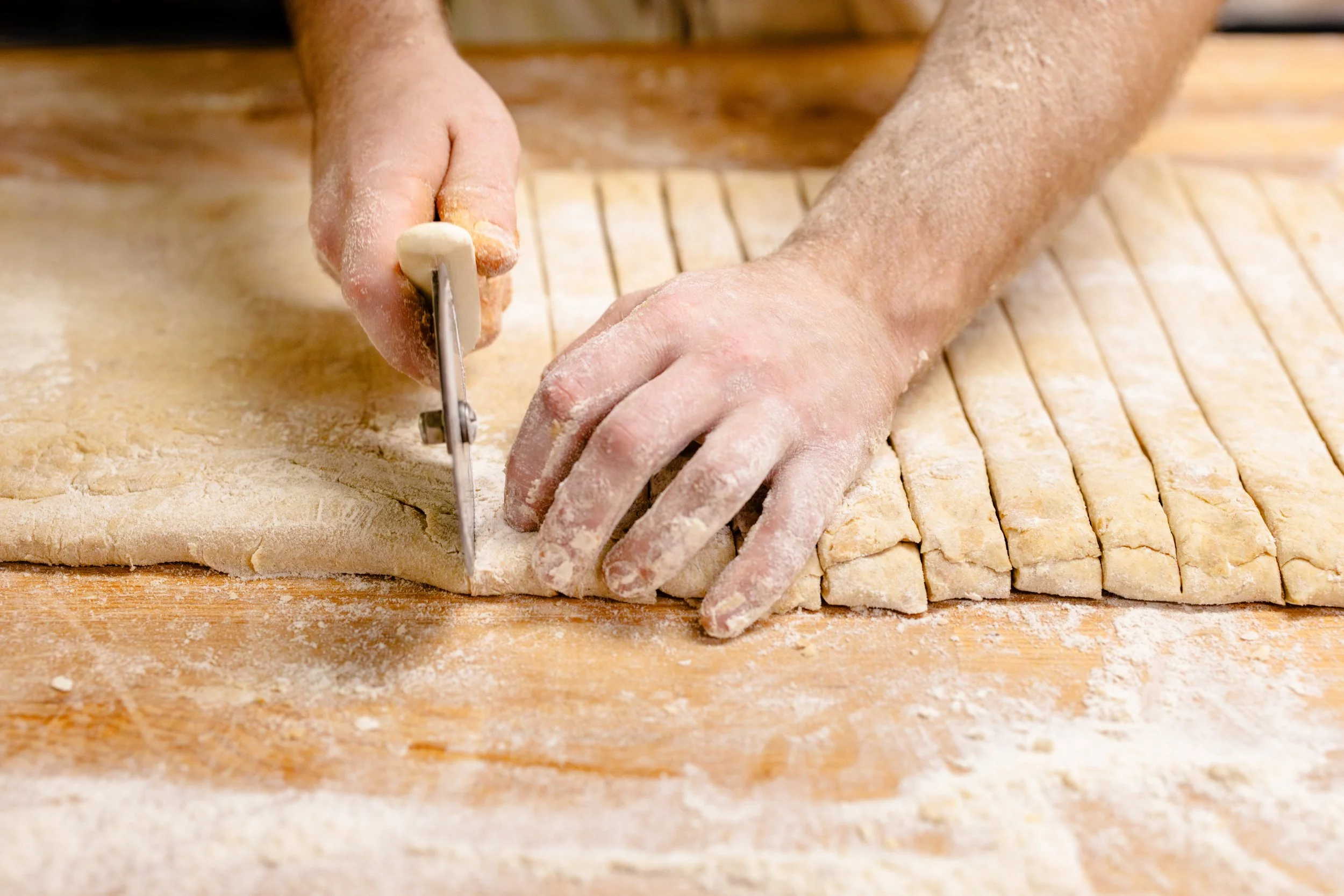 Person slicing rolled out dough into strips on a floured wooden surface.