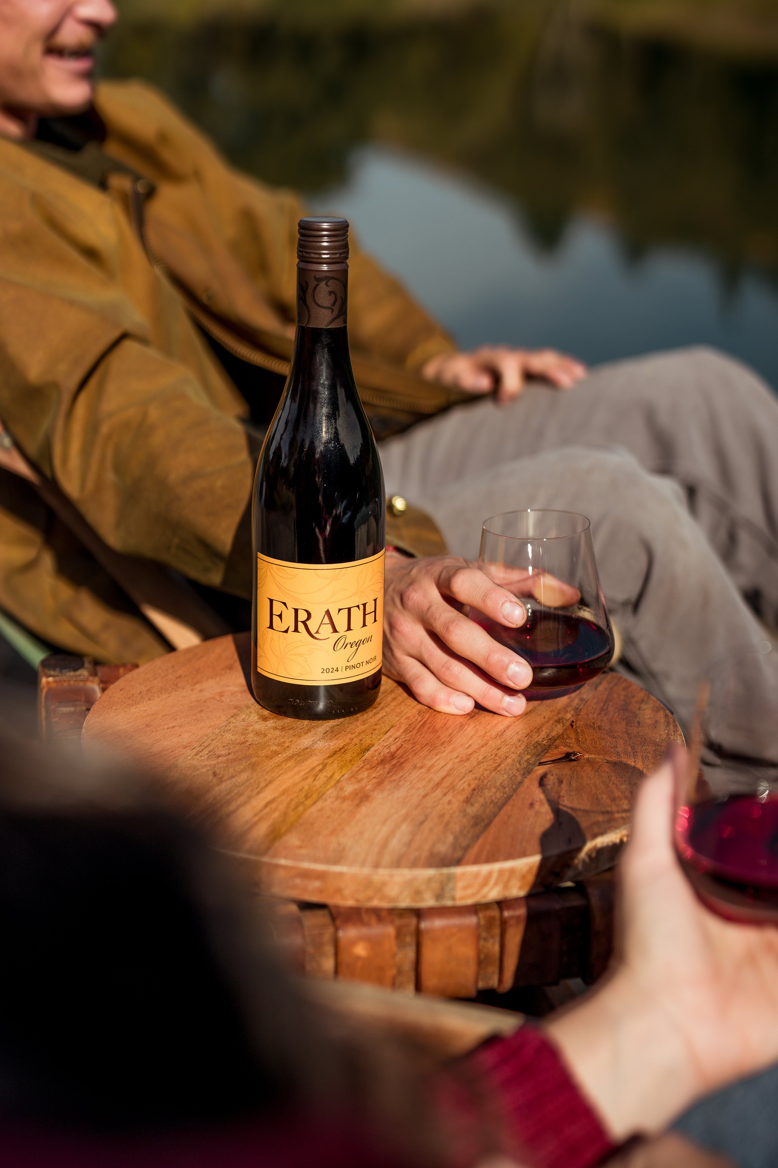 A person holding a glass of red wine next to a bottle labeled 'ERATH Oregon' on a wooden table outdoors near a body of water.
