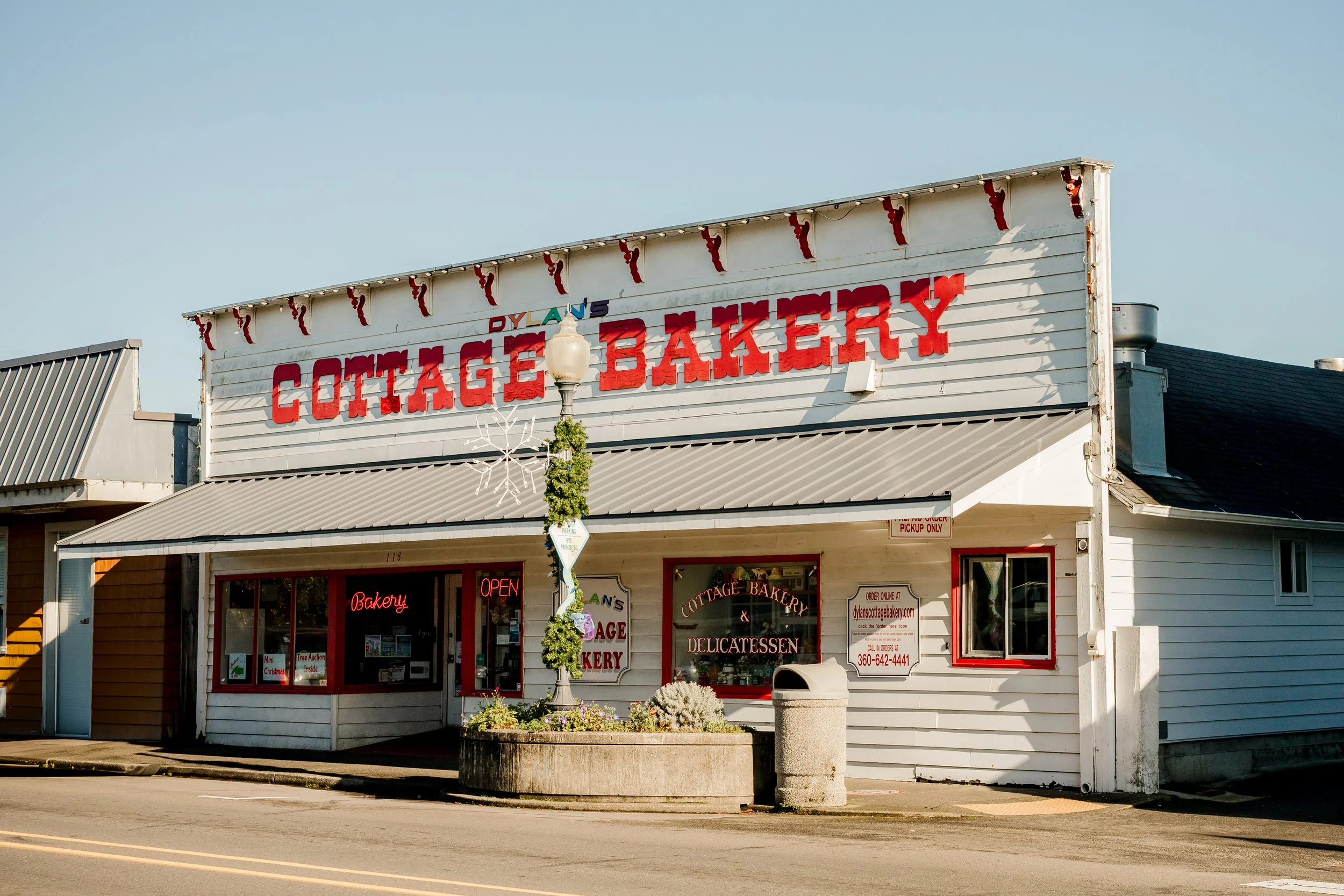 A storefront with a sign reading "Dylan's Cottage Bakery" in red letters, featuring a small patio with a lamppost decorated with greenery and flowers, and windows displaying baked goods.