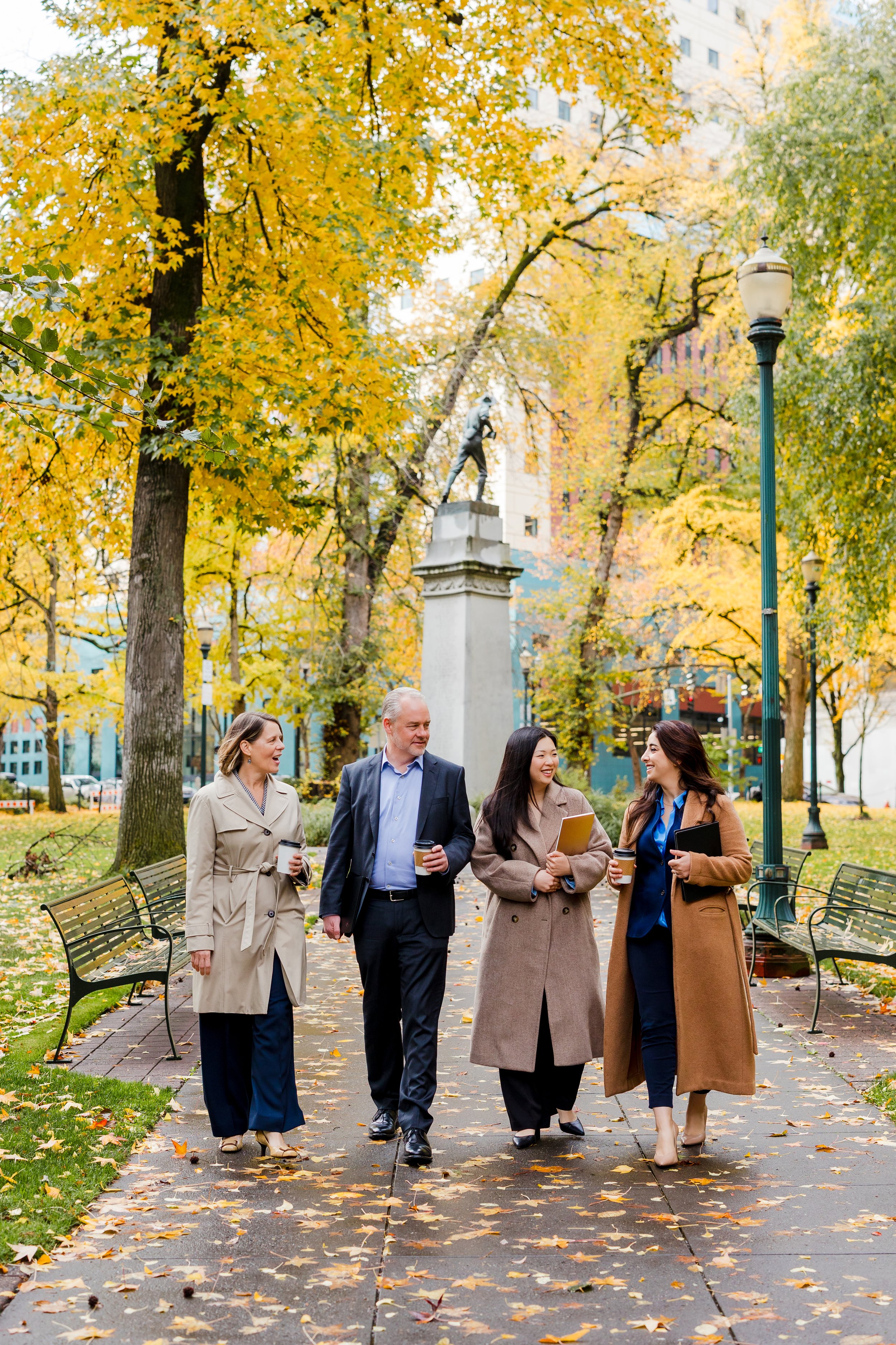 Four professionally dressed people walking and talking in a park during fall, holding coffee cups, with trees and a statue in the background.