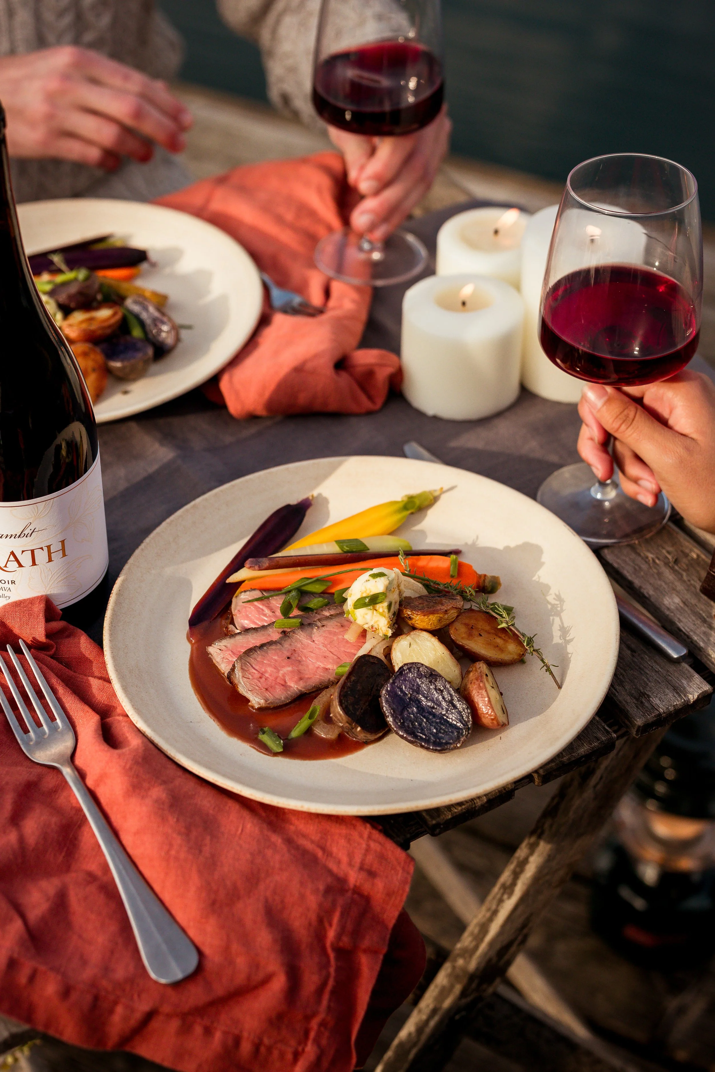 A dining scene with two plates of food, glasses of red wine, lit candles, and a bottle of wine on a rustic wooden table.