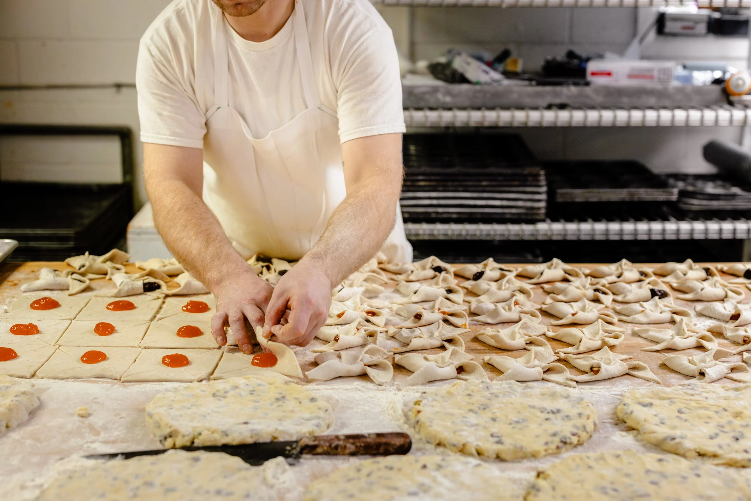 Baker preparing small square pastries with cherry filling and cherry topping on a floured work surface in a bakery kitchen.