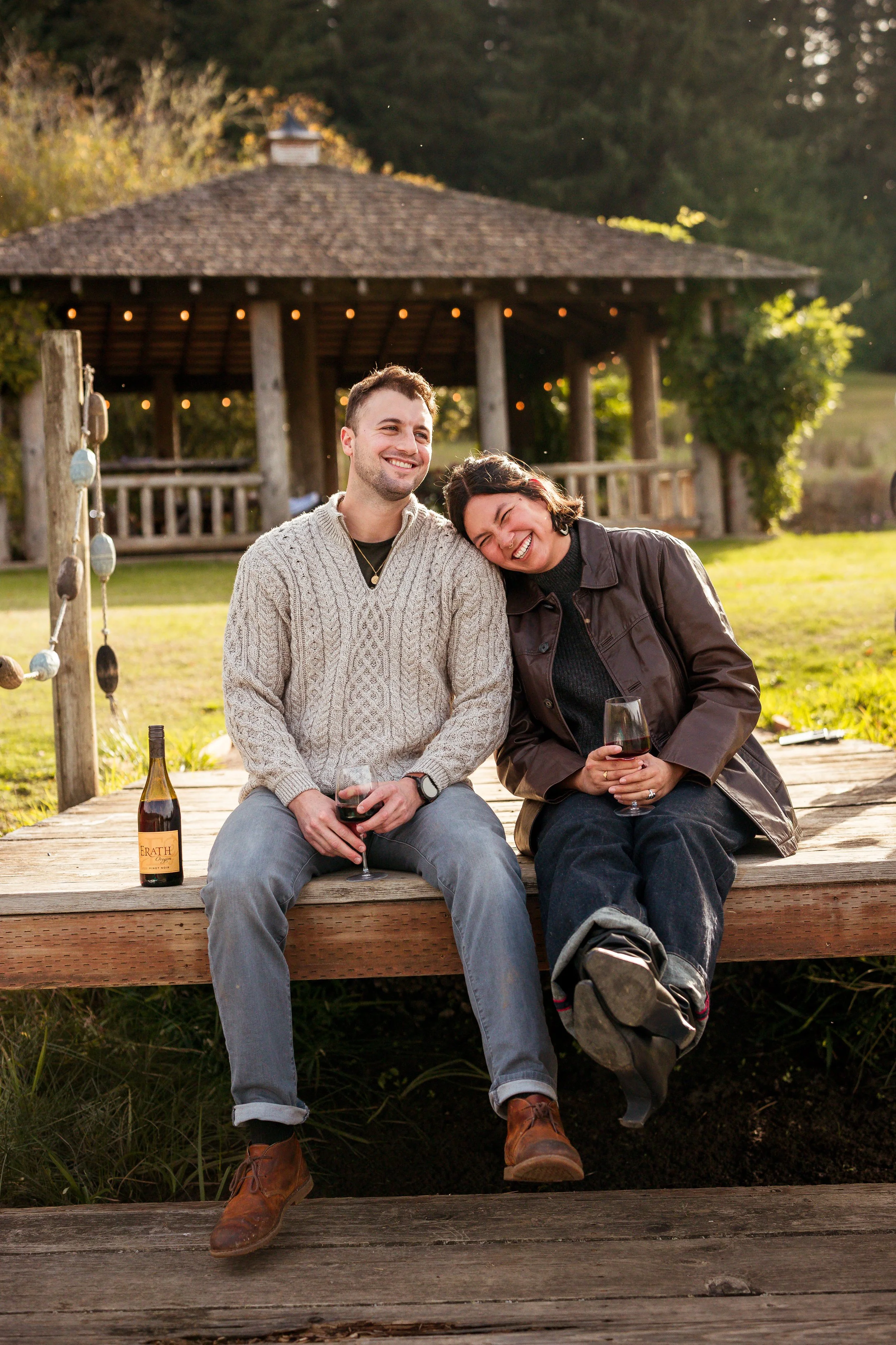 A man and woman sitting on a wooden dock, smiling and laughing, holding glasses of red wine, with a bottle of wine beside them, in front of a gazebo surrounded by trees and greenery in a park.