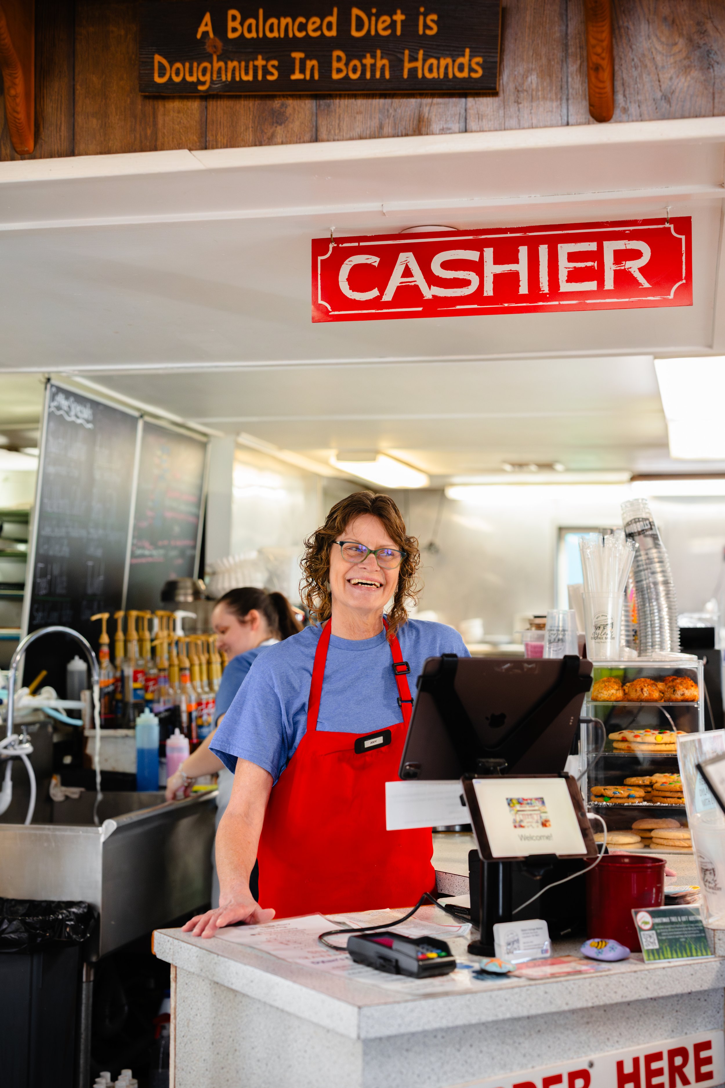 Female cashier with curly hair and glasses smiling at the checkout counter in a bakery or café with donuts and baked goods behind her.