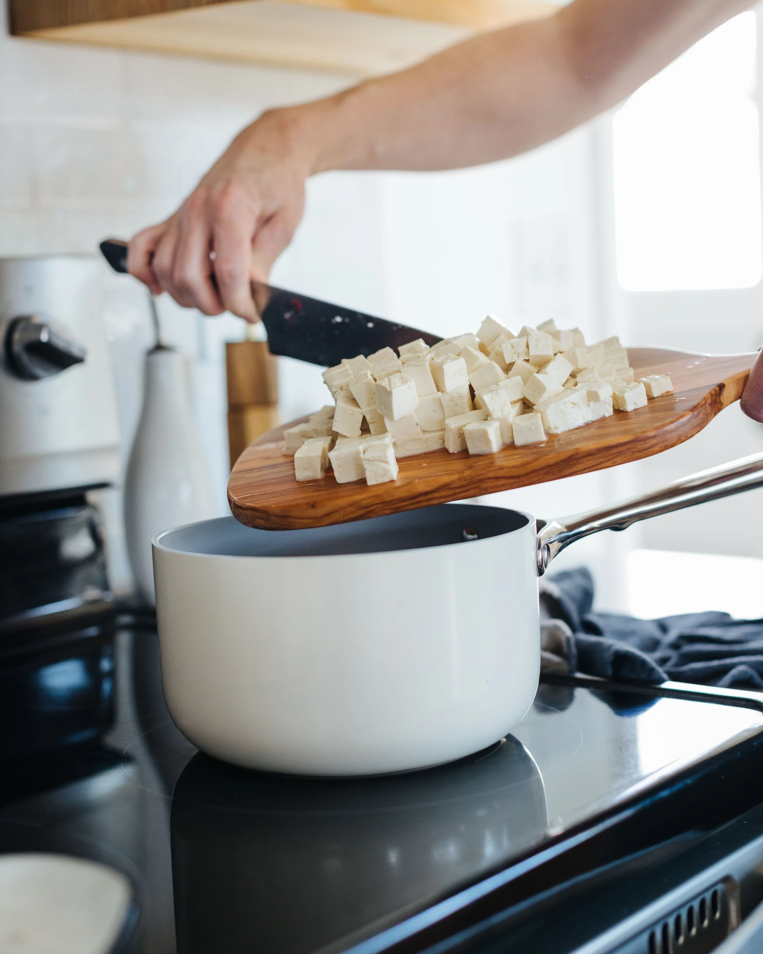 Person chopping tofu on a wooden cutting board over a white pot on a stove.