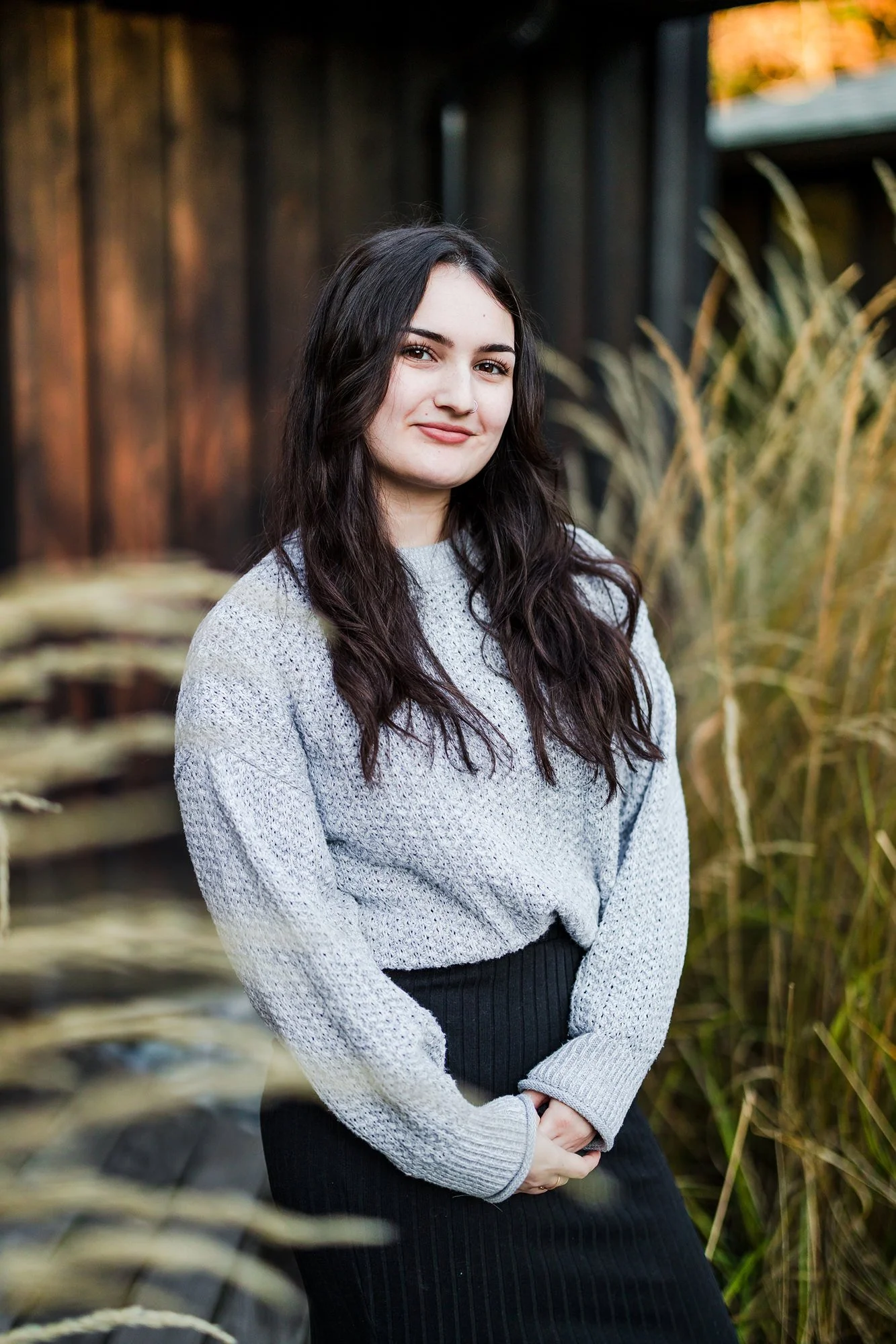 A young woman with long dark wavy hair, wearing a light gray sweater and a black skirt, standing outdoors next to tall grass and a wooden fence