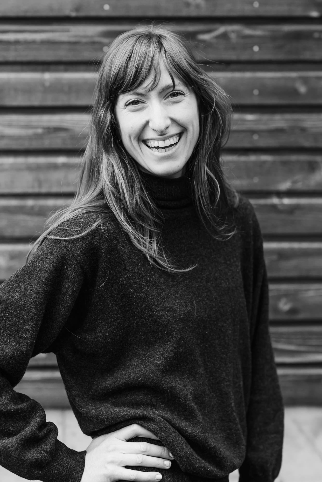 Black and white portrait of a young woman with long, wavy hair smiling, wearing a dark turtleneck sweater, standing in front of wooden planks.