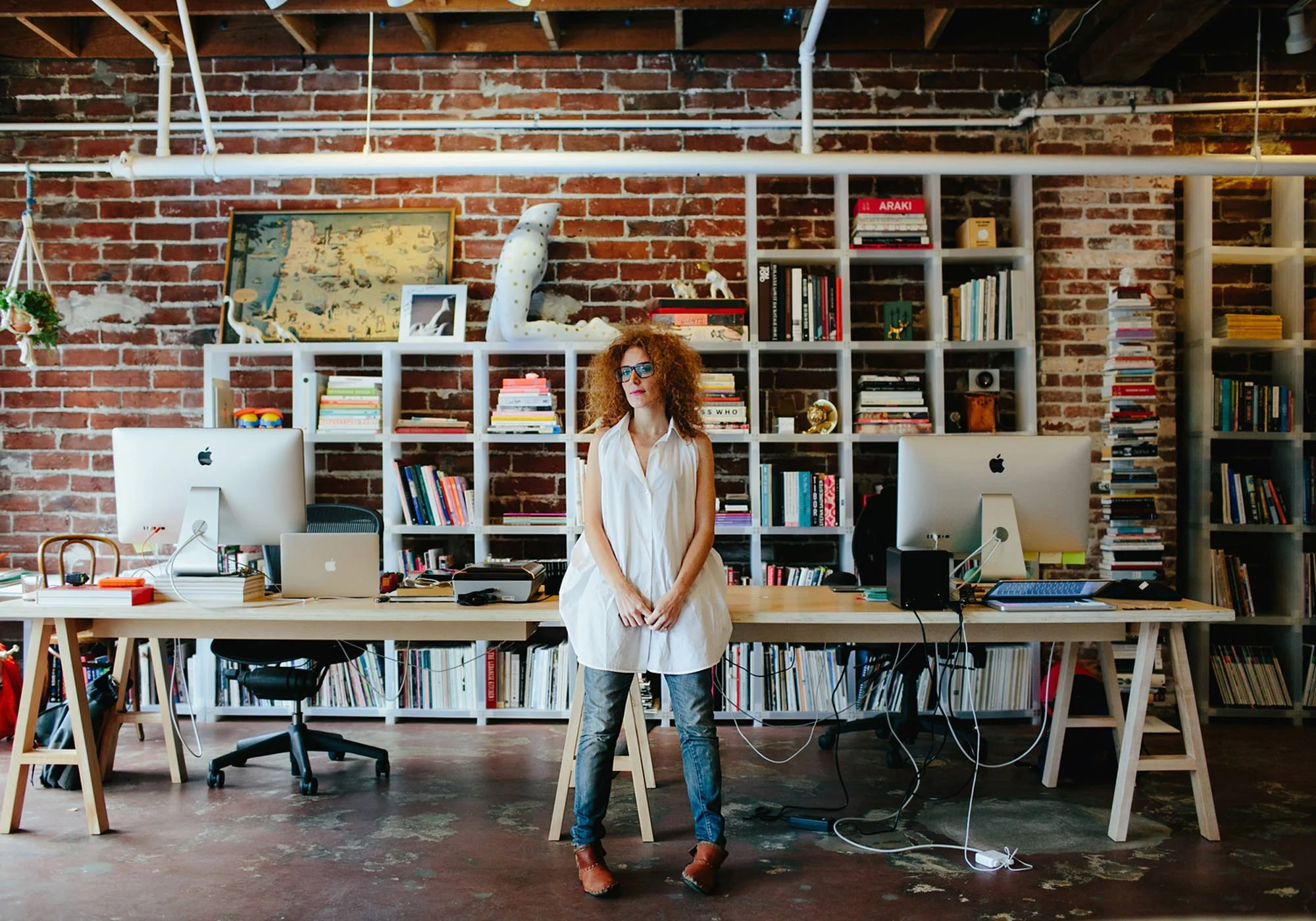 Woman standing in front of a large wooden desk in a modern office with exposed brick walls and white bookshelves filled with books and decorative items.