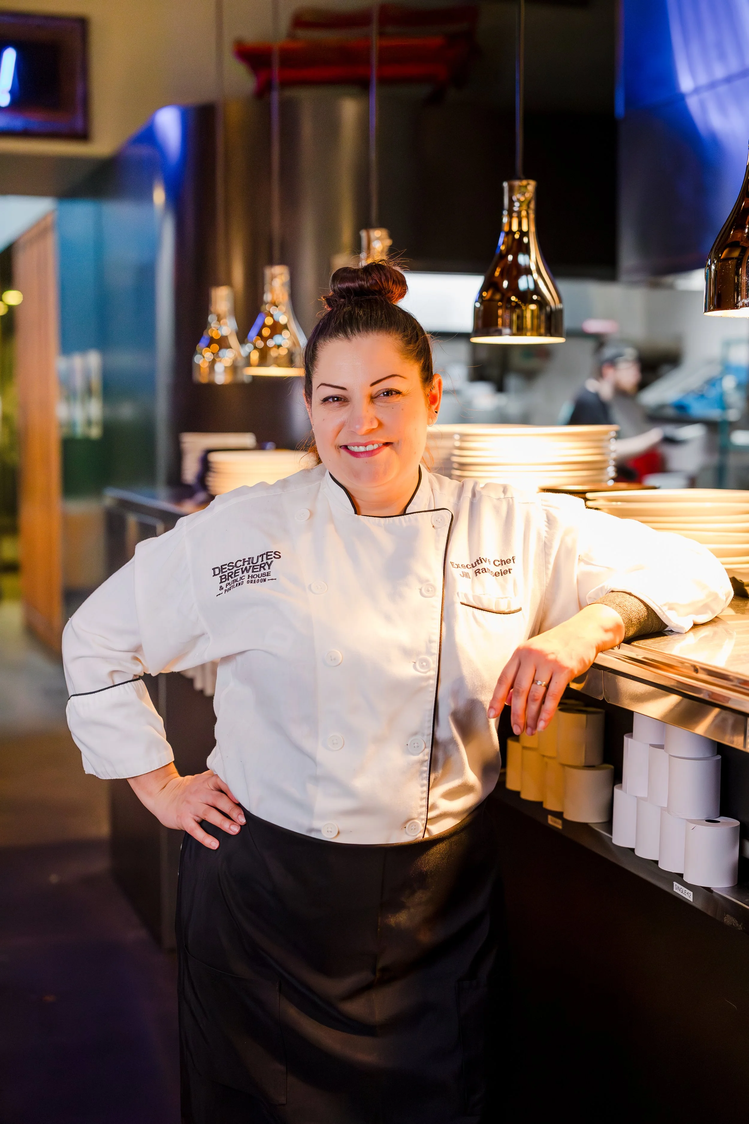A smiling female chef in a white chef's coat in a restaurant kitchen leaning against a stainless steel counter with plates behind her.