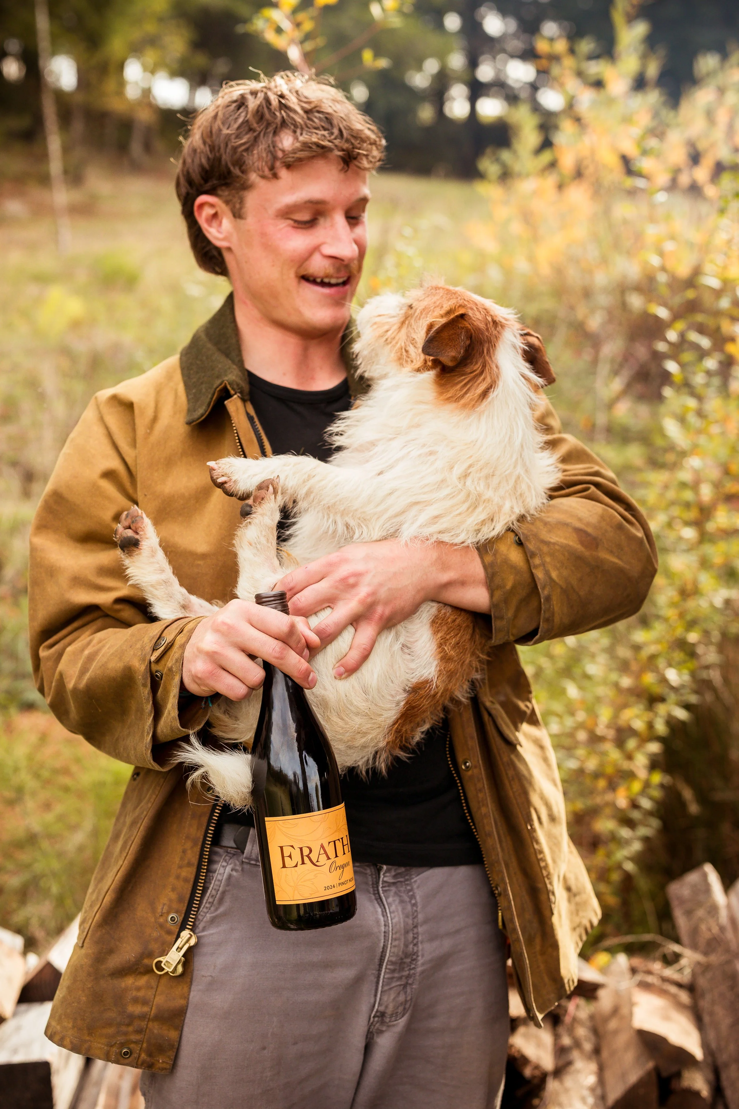 A man holding a small dog and a bottle of wine outdoors in a wooded area.