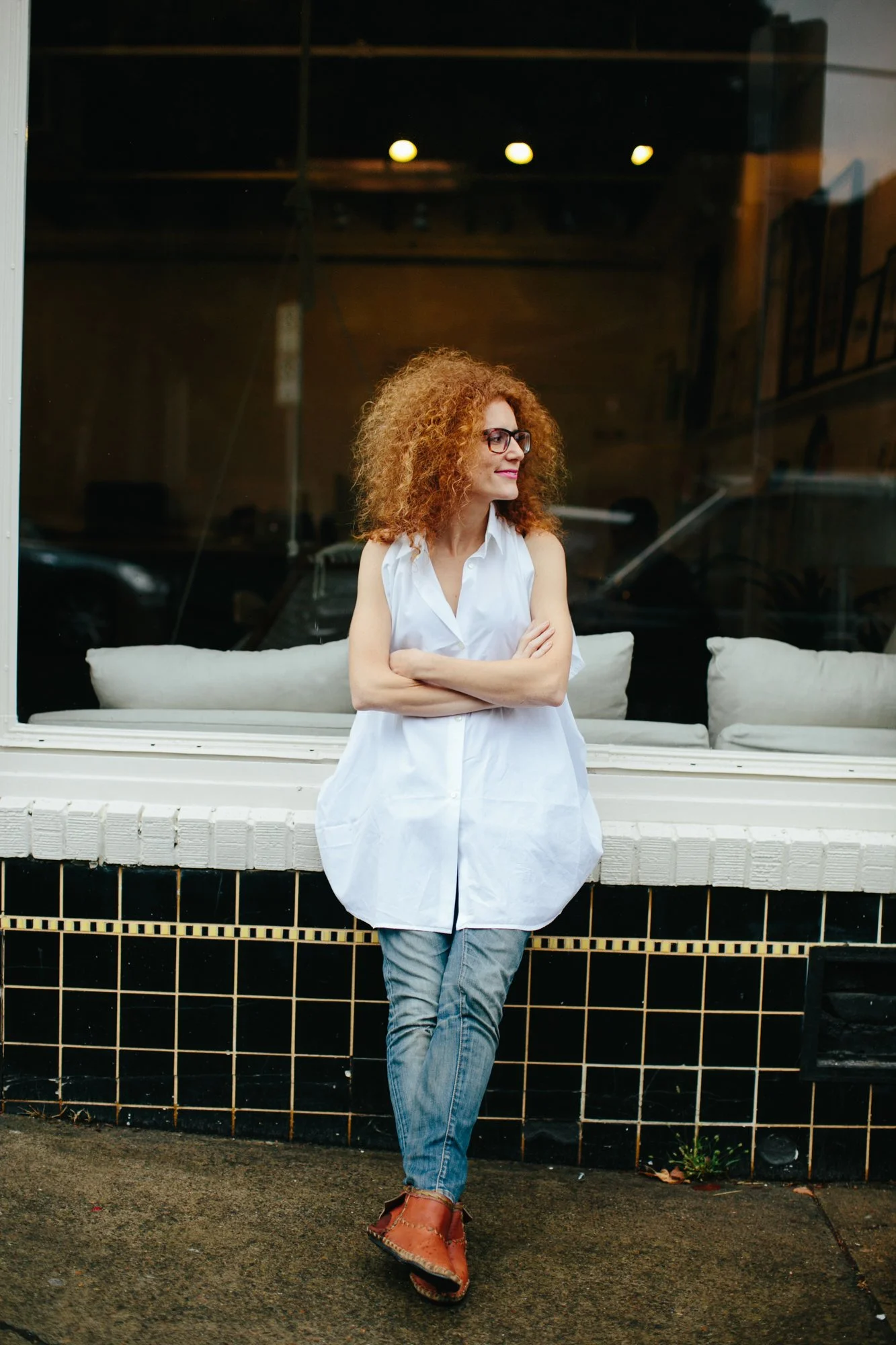 A woman with curly red hair, glasses, and a white sleeveless shirt standing outside a cafe or shop window with arms crossed, looking to the side.