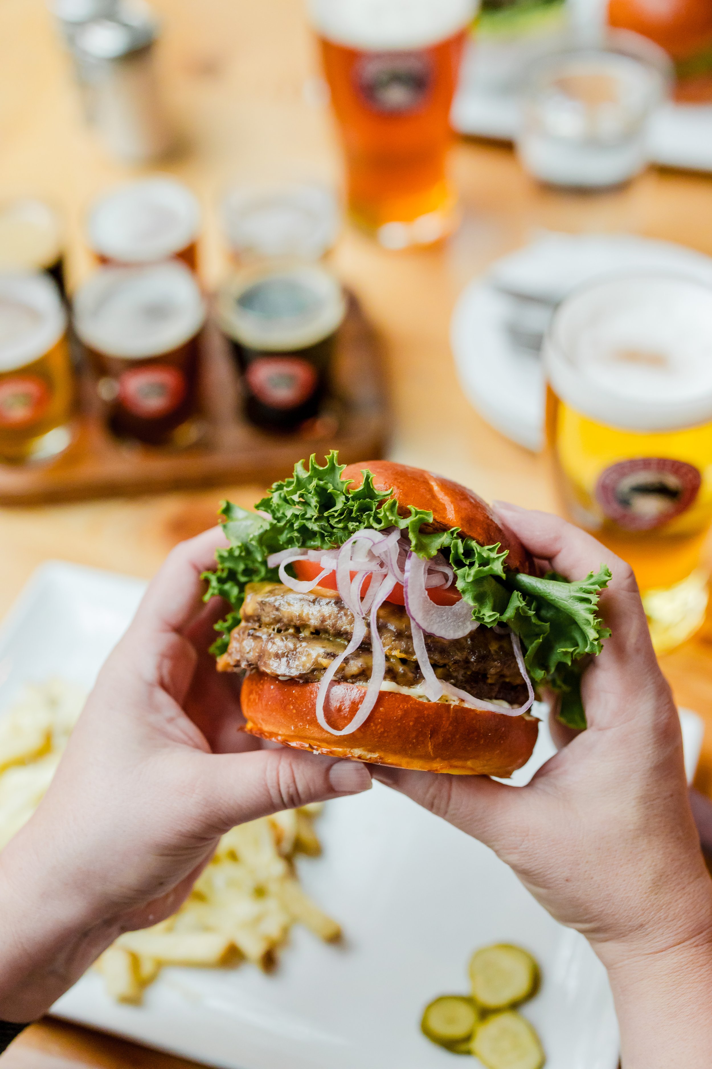 Person holding a hamburger with lettuce, tomato, onions, and a beef patty at a table with beers and condiments in the background.