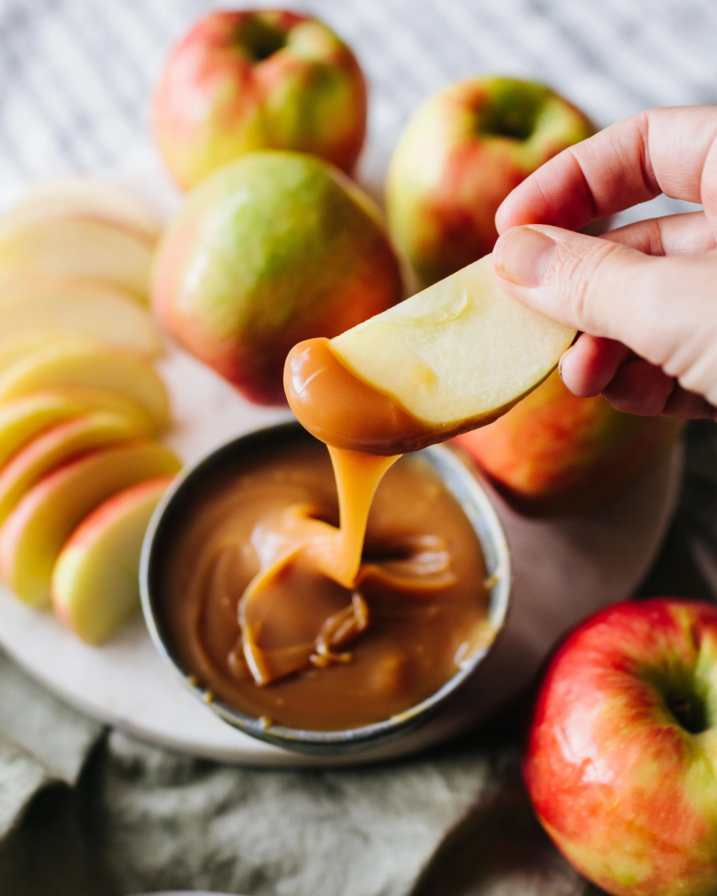 A person dipping a slice of apple into caramel sauce with apples and caramel on a plate in the background.