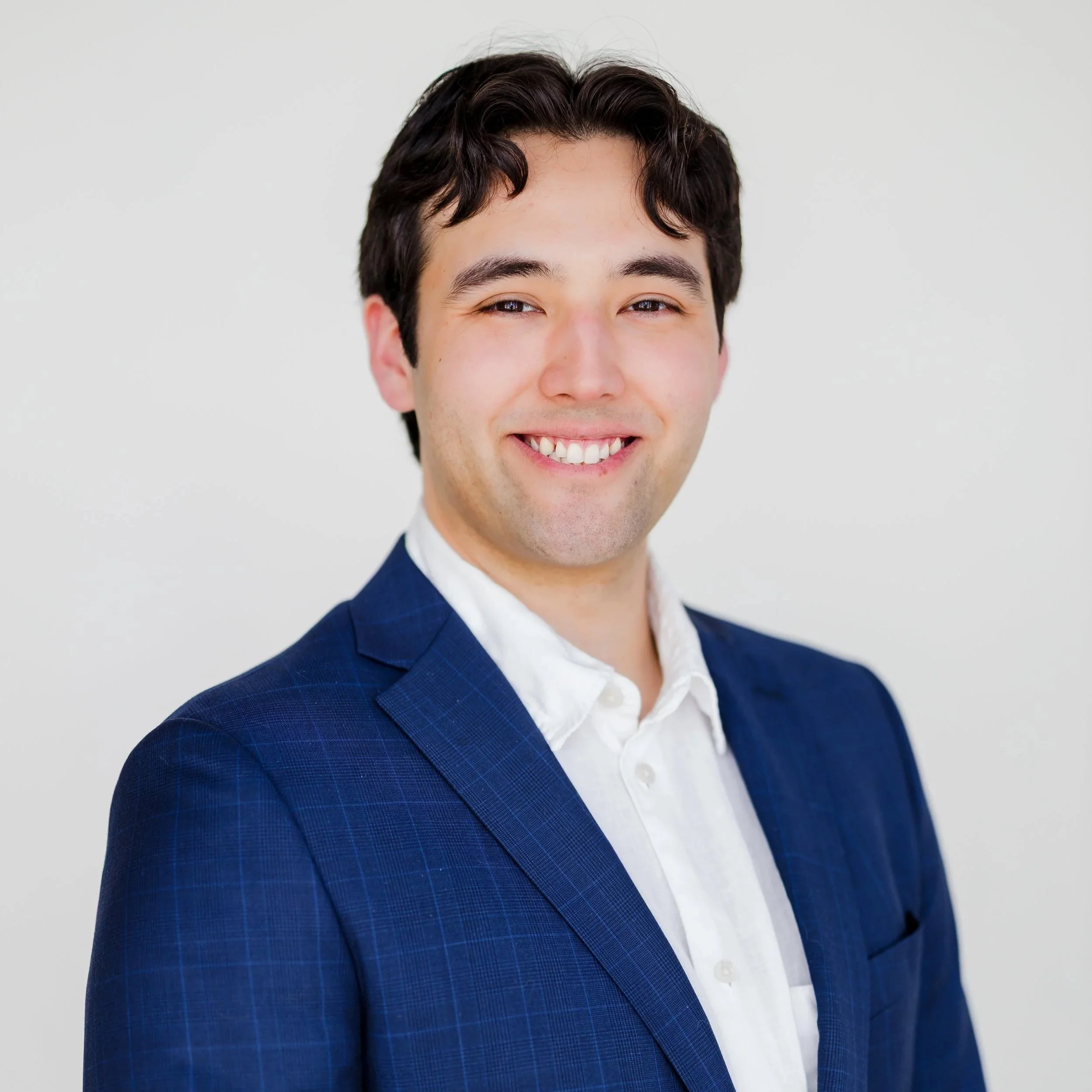 Portrait of a smiling young man with dark hair, wearing a blue checkered suit jacket and white shirt, standing against a plain light background.