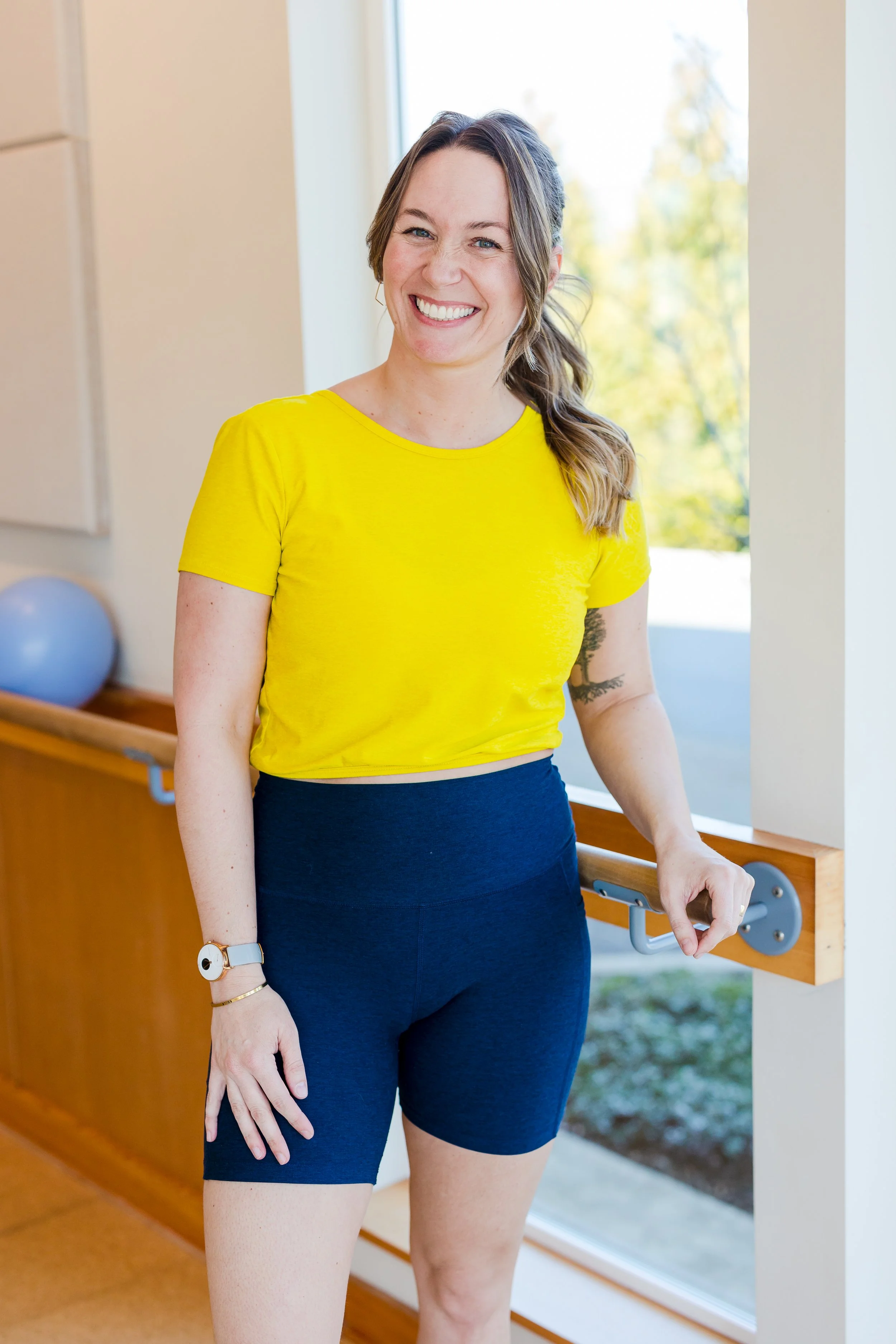 A woman with brown hair tied in a ponytail, wearing a yellow t-shirt and navy shorts, standing near a window in a bright room, smiling at the camera.