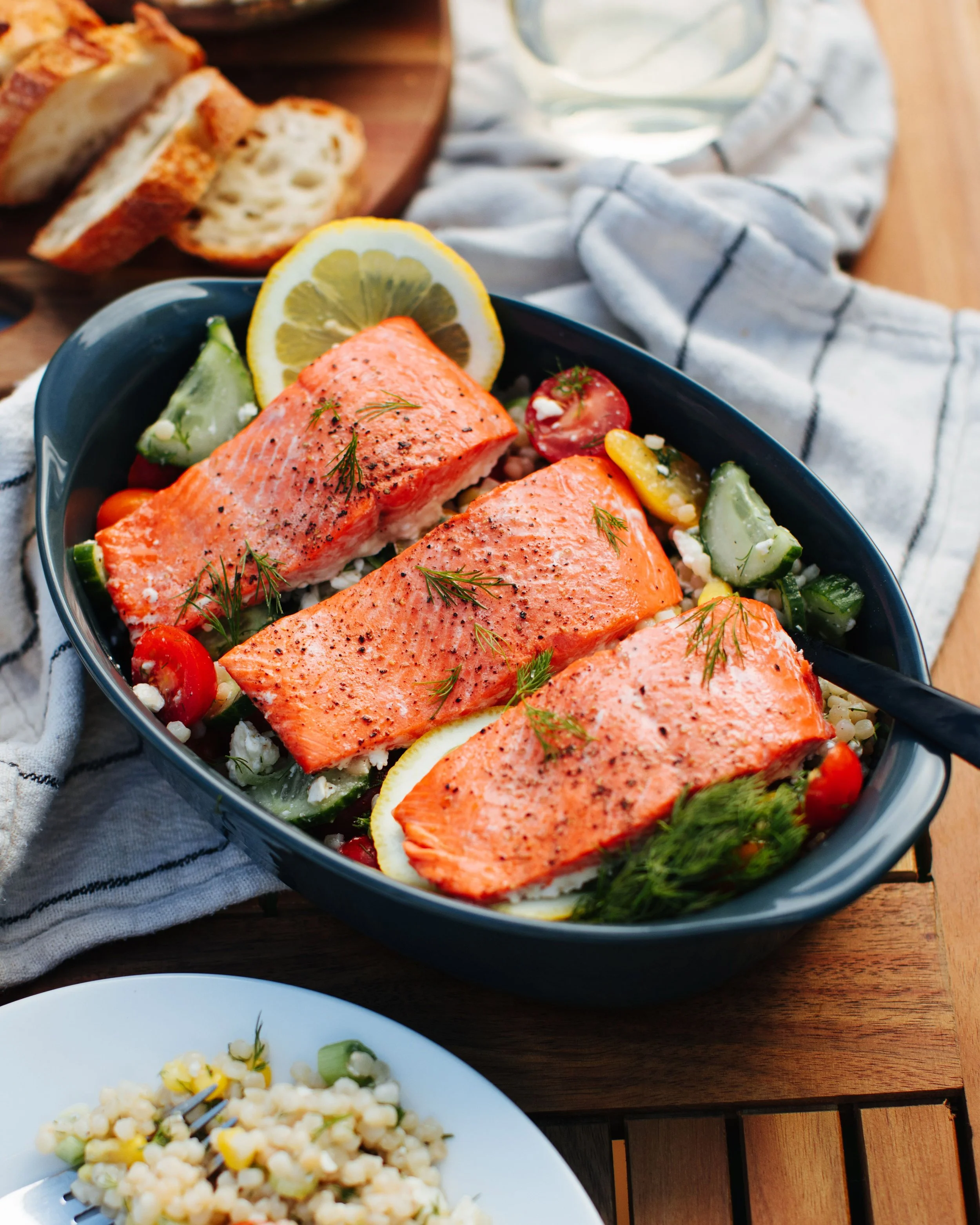 Baked salmon fillets garnished with fresh herbs and lemon slices in a black dish, with cucumber, cherry tomatoes, and herbs around it, on a wooden table with a blue striped cloth.