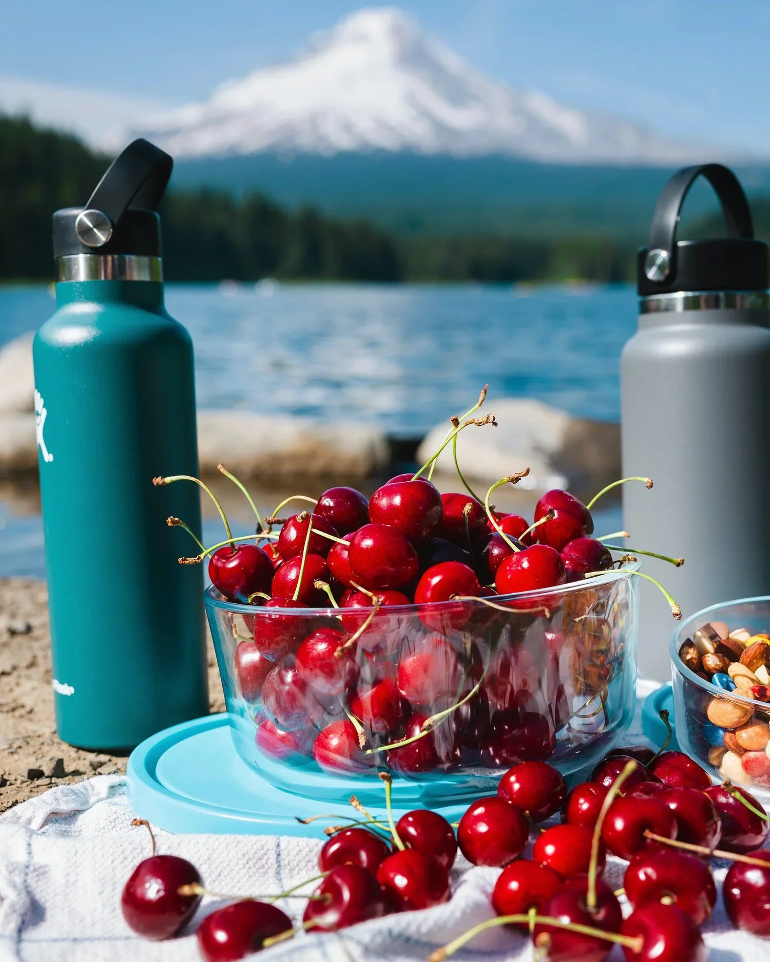 A picnic setup by a lake with a background of snow-capped mountain, featuring a glass bowl of fresh cherries, two water bottles, and a bowl of mixed nuts on a white cloth.