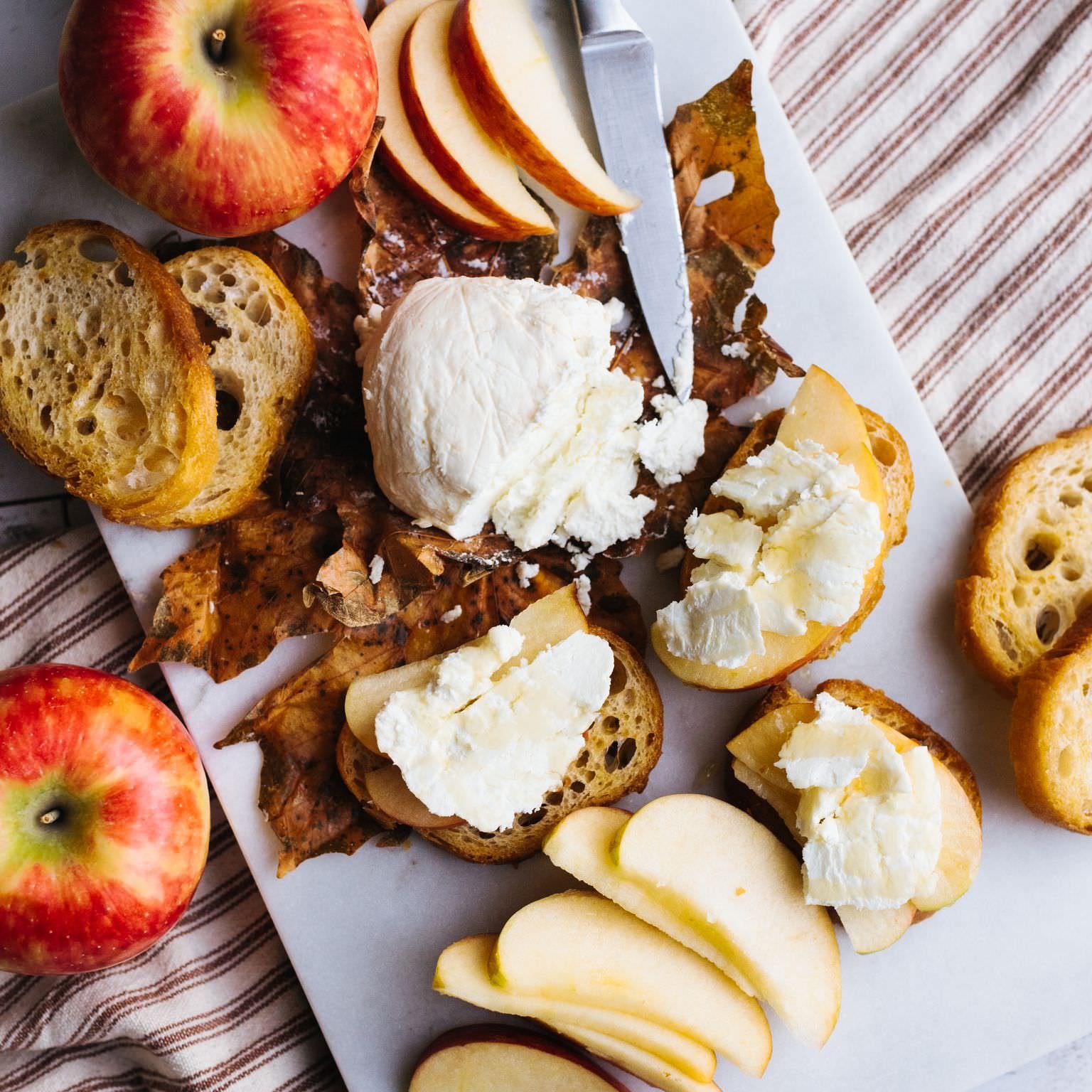 A cheese platter with apples, bread, and a knife on a white tray on striped cloth.