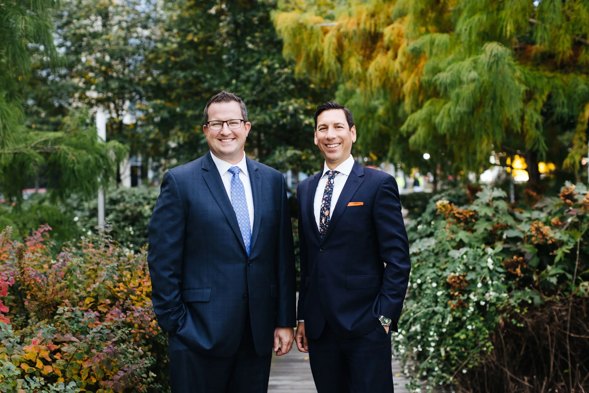 Two men in suits standing outdoors on a pathway surrounded by greenery and fall foliage, smiling at the camera.