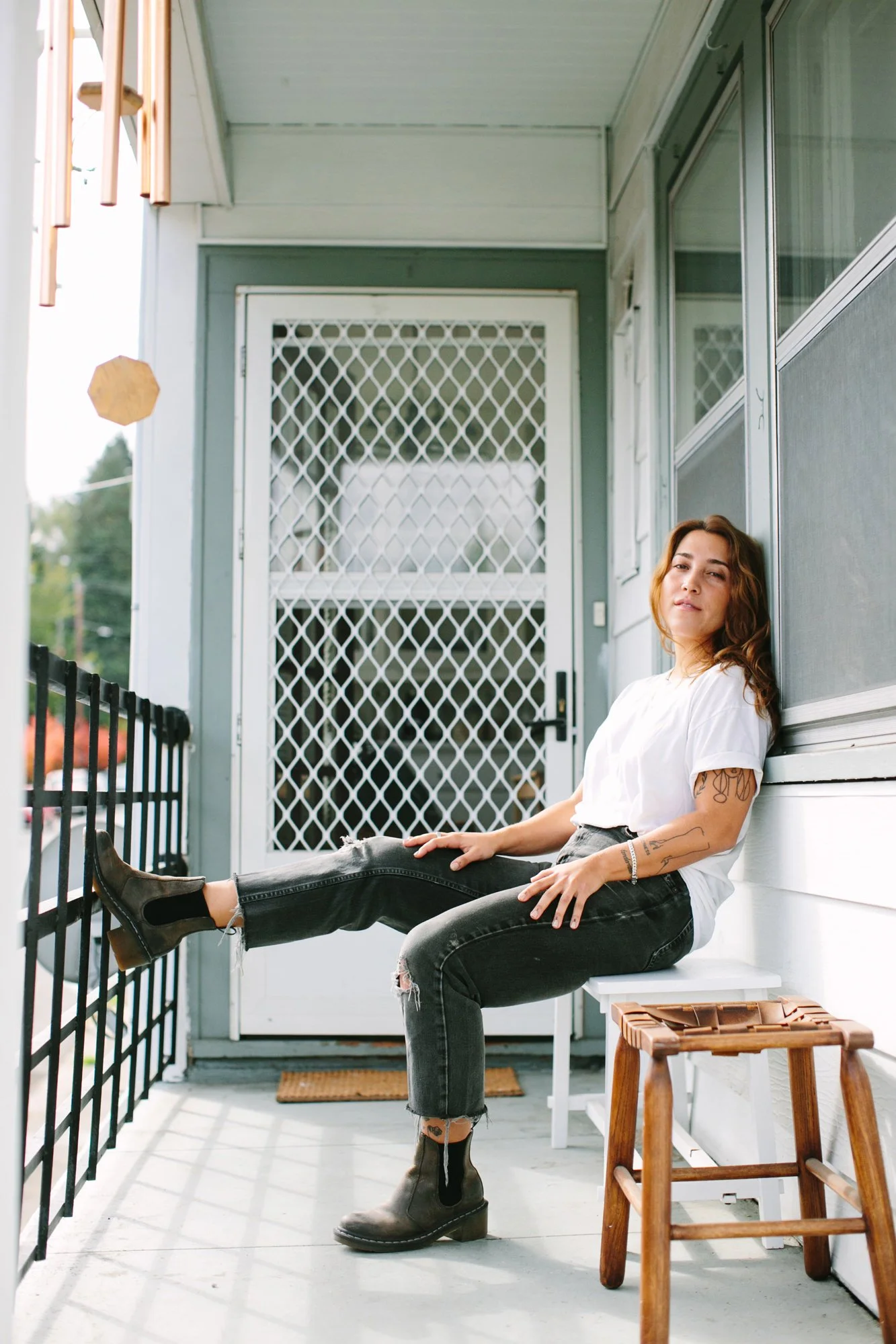 A woman sitting on the floor of a porch, leaning against the wall, with one leg extended and resting on the porch railing. She has brown wavy hair, wears a white t-shirt, black ripped jeans, and ankle boots, and is looking toward the camera with a re