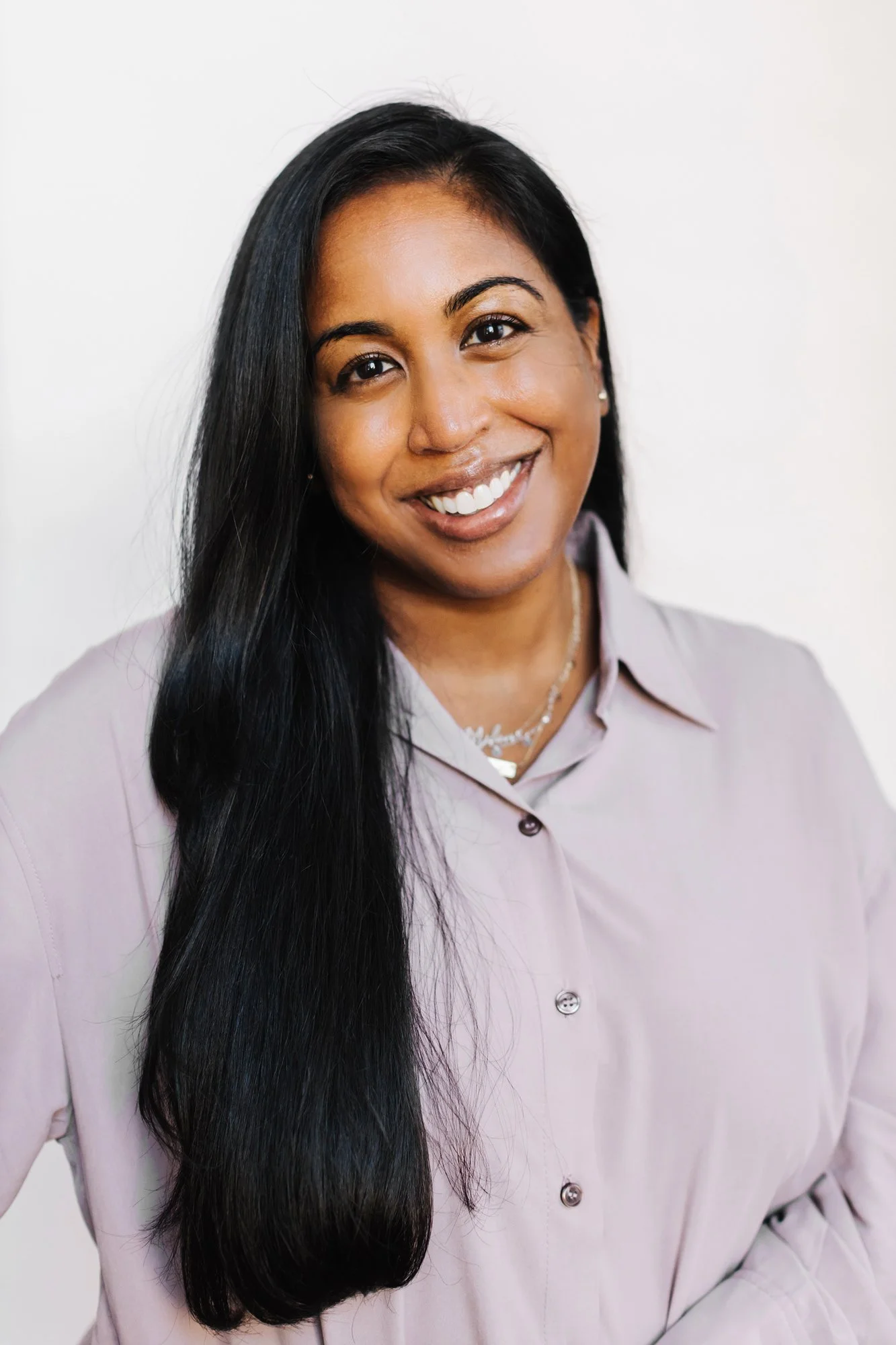 A smiling woman with long black hair wearing a light purple button-up shirt and silver jewelry, standing against a white background.