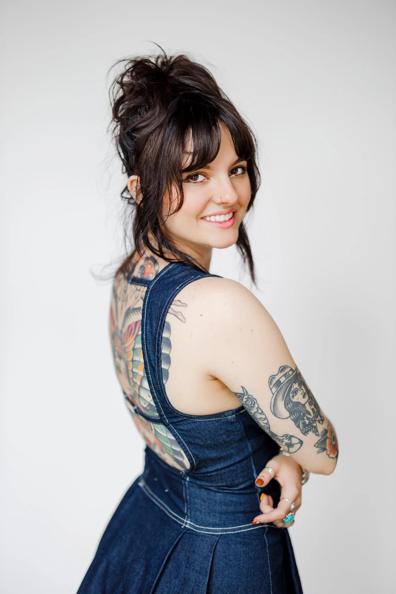 A woman with dark hair tied in a loose bun, smiling and looking at the camera, wearing a sleeveless denim dress with colorful tattoos on her arm and back, standing against a plain white background.