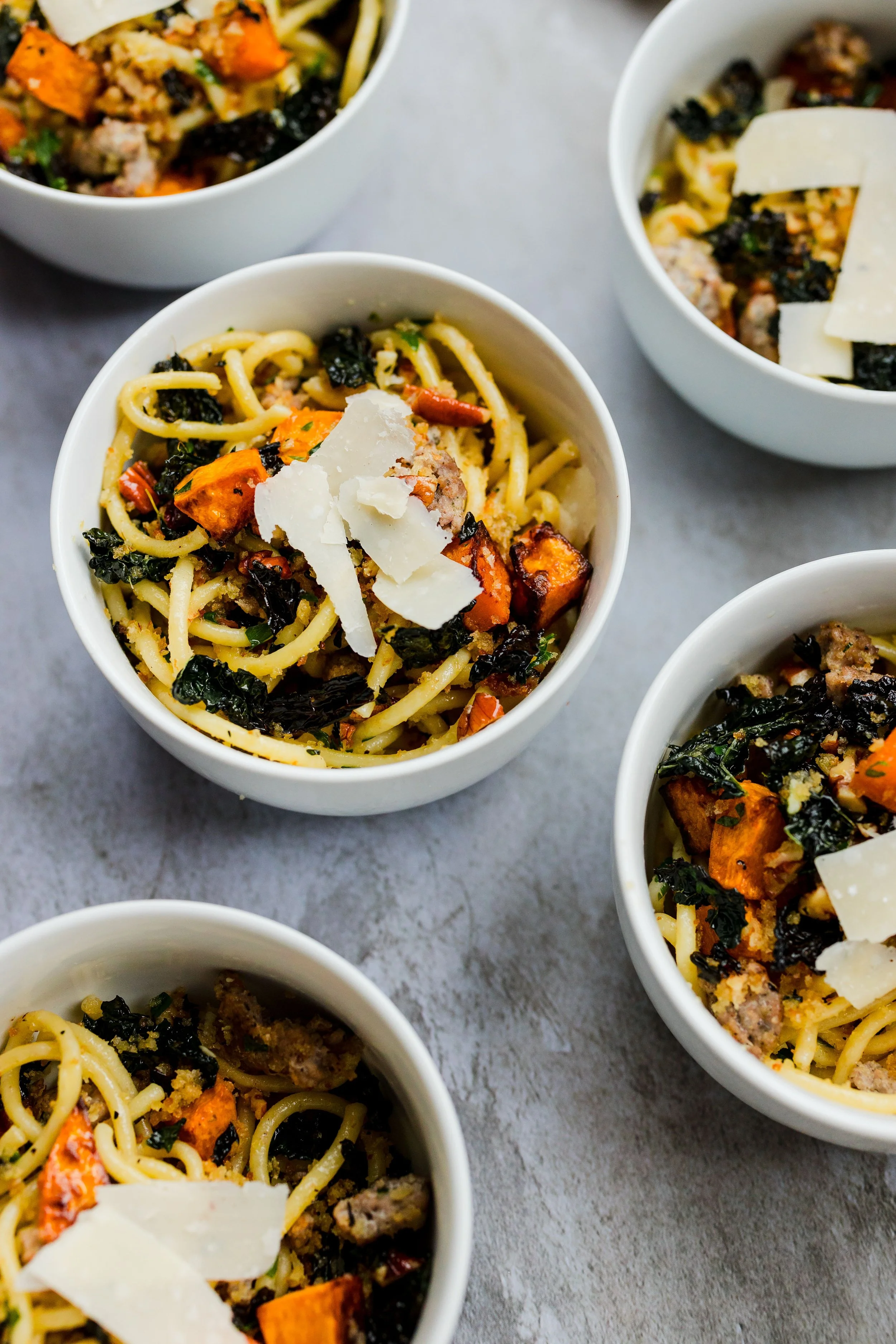 Four white bowls of spaghetti with chopped vegetables, ground meat, kale, topped with shaved cheese, on a gray surface.