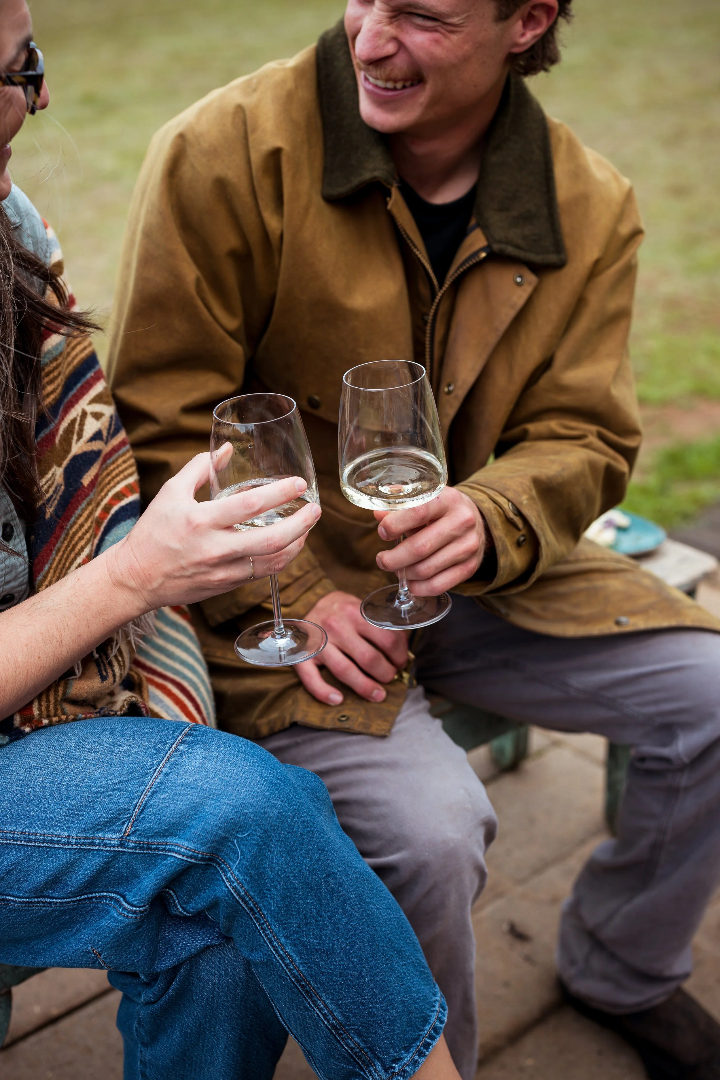 People sitting outdoors, holding glasses of white wine, engaged in conversation, wearing casual autumn clothing.