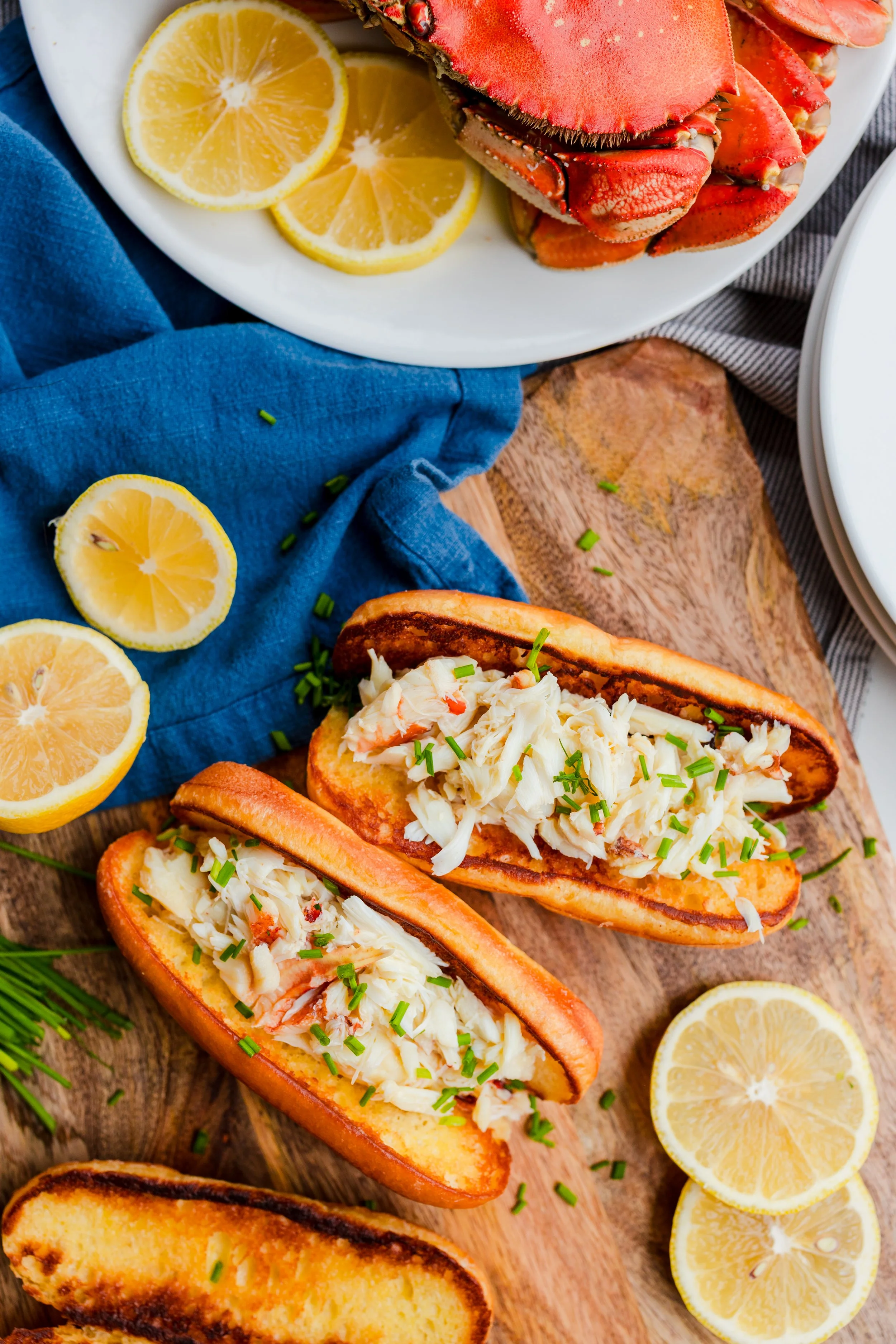 Lobster, lemon slices, clam roll sandwiches with chopped chives on a wooden cutting board, under a blue cloth.