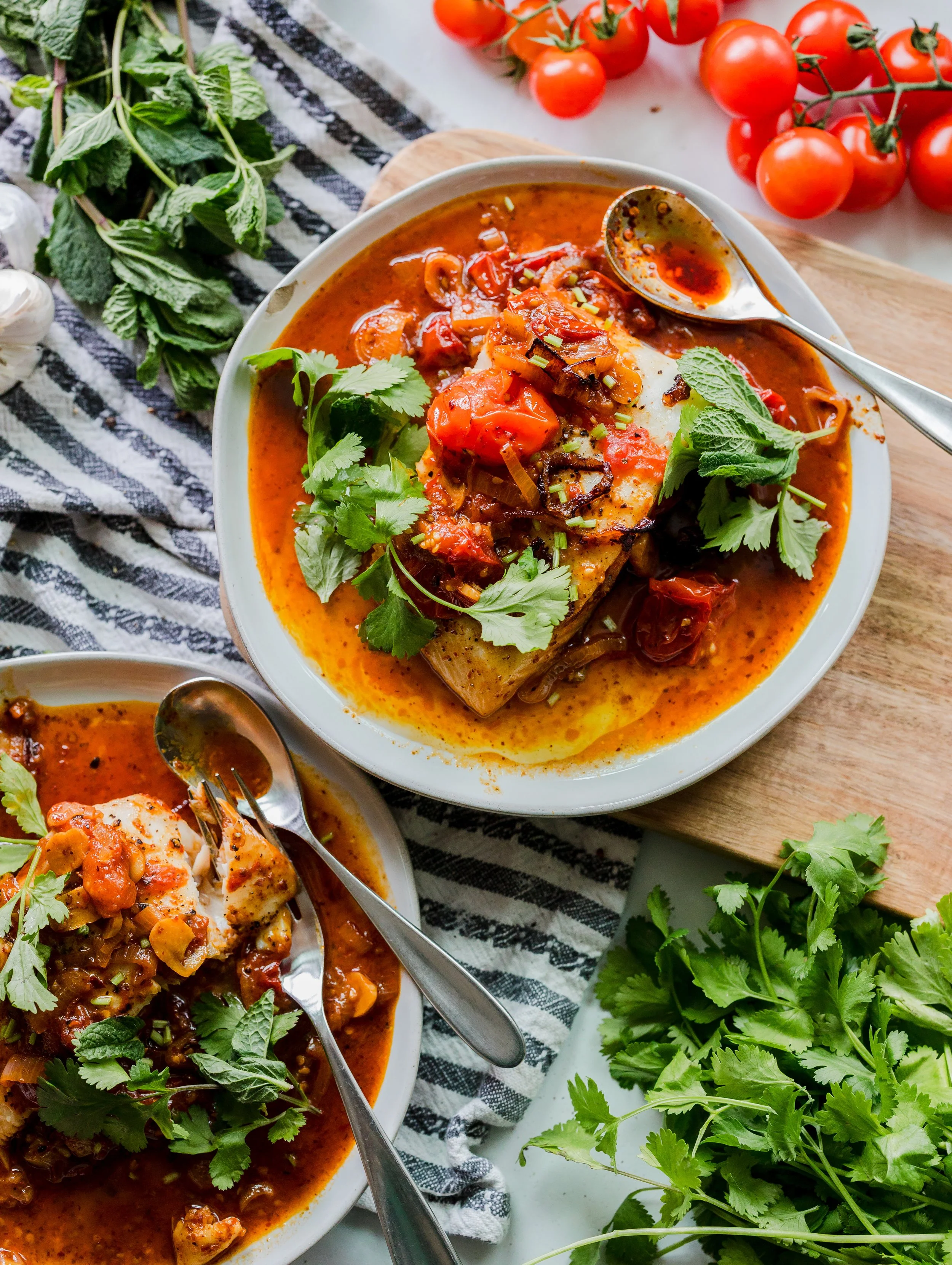 Two bowls of fish stew garnished with cilantro, with cherry tomatoes, garlic, and fresh herbs around them.