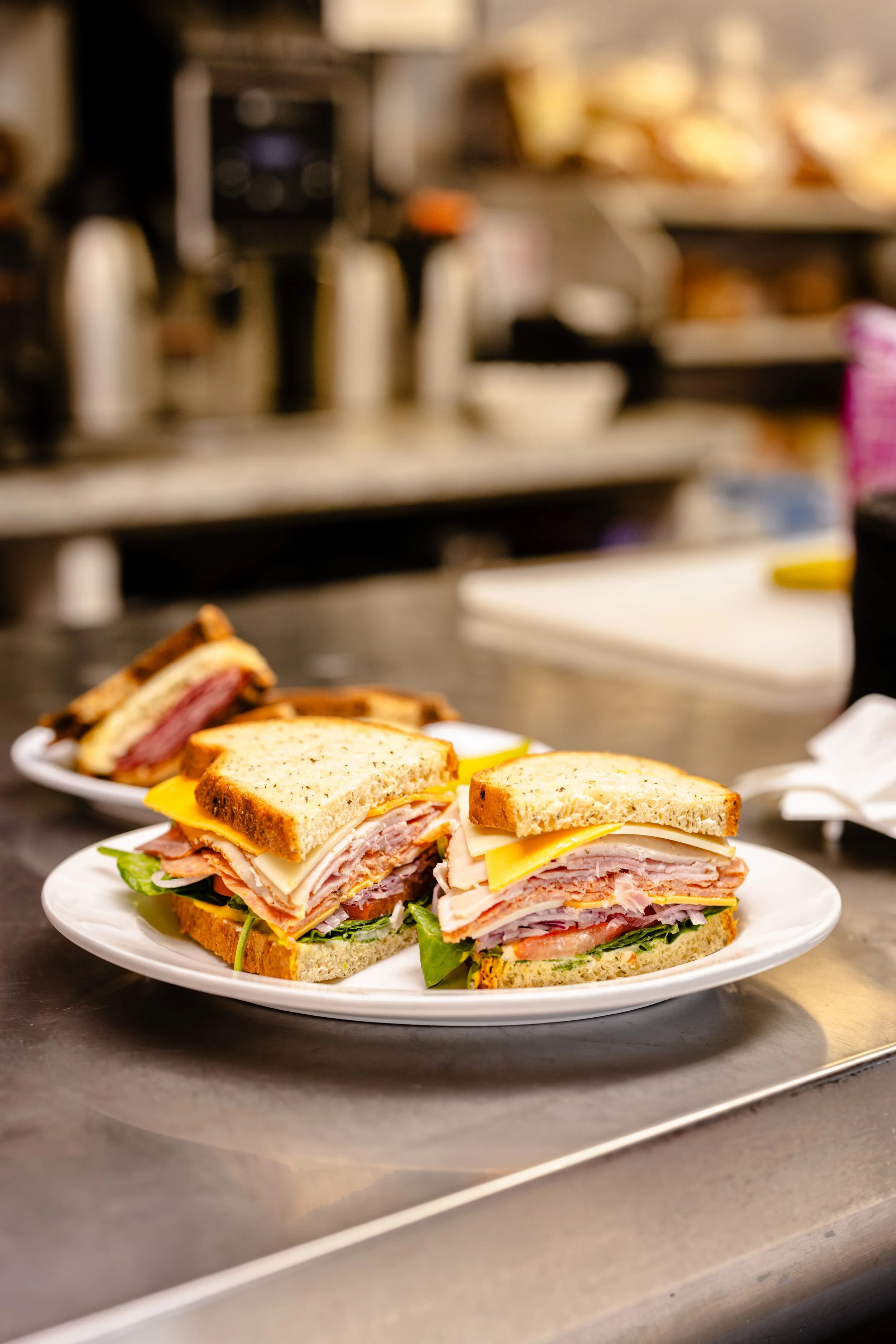 Plates of toasted sandwiches with layers of turkey, cheese, ham, lettuce, and tomato on a stainless steel counter in a kitchen.