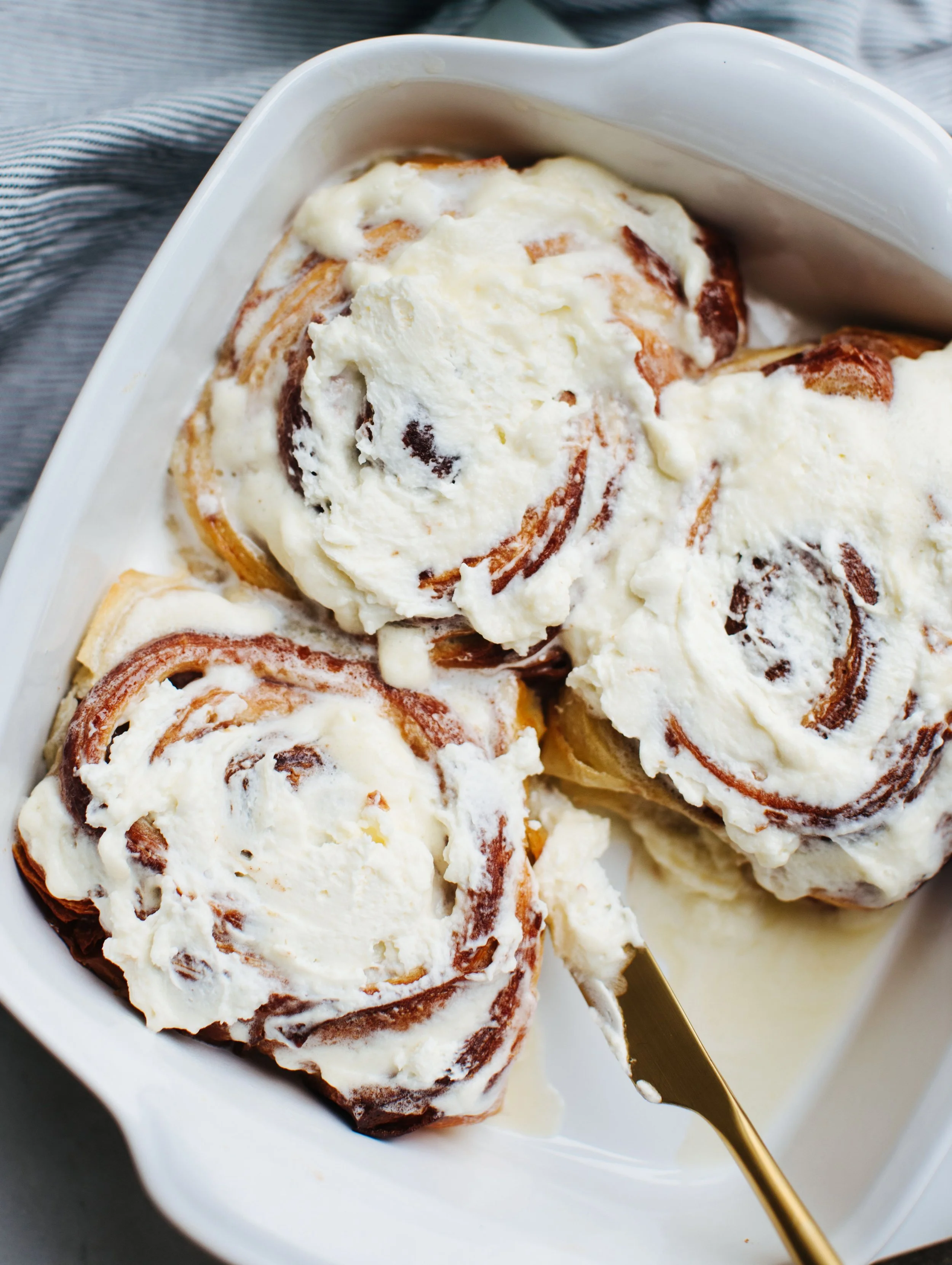 Close-up of three cinnamon rolls with icing in a white baking dish, with a gold serving utensil.