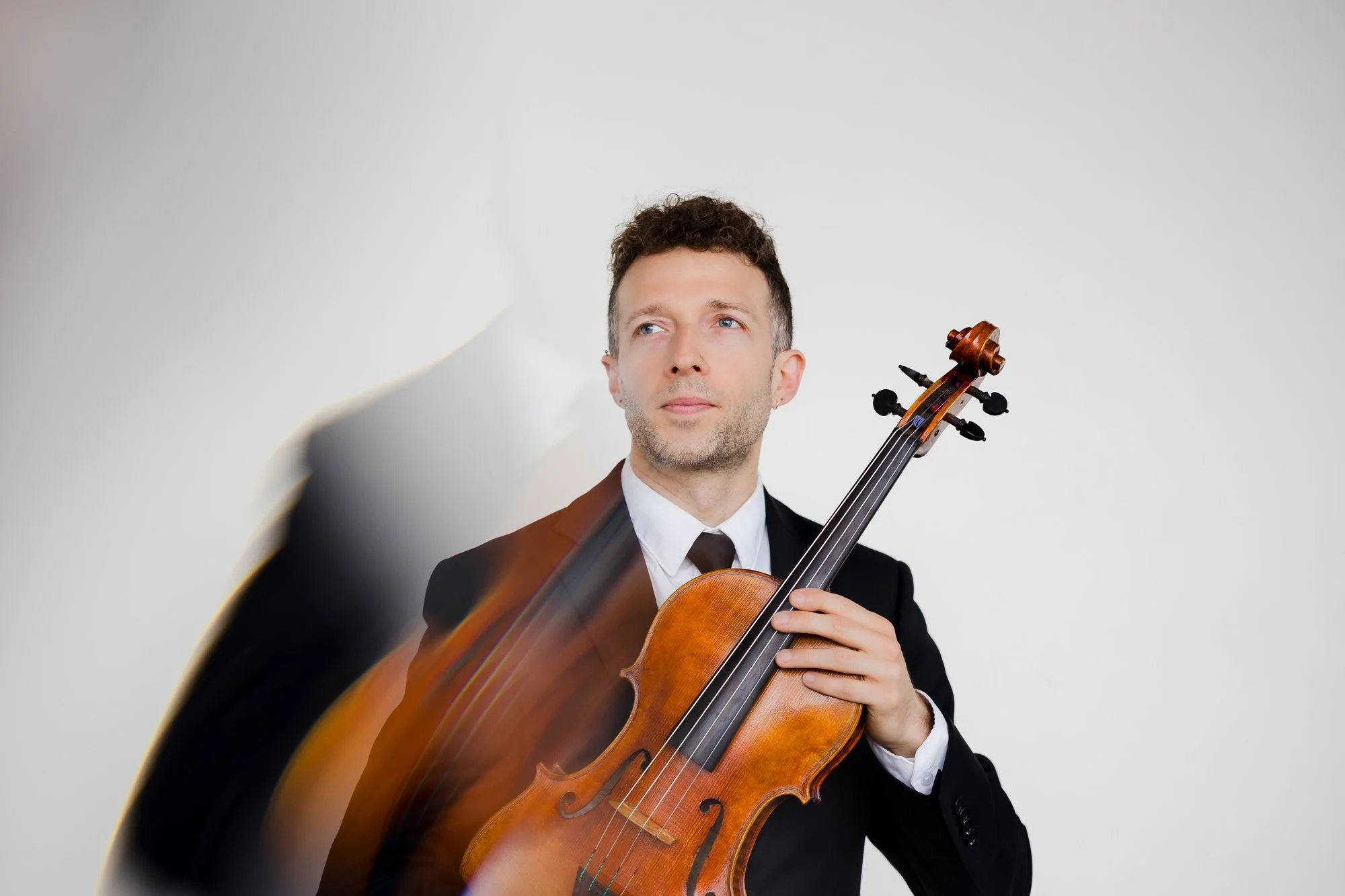 A man in a black suit holding a violin, looking thoughtfully to the side, with a neutral background.