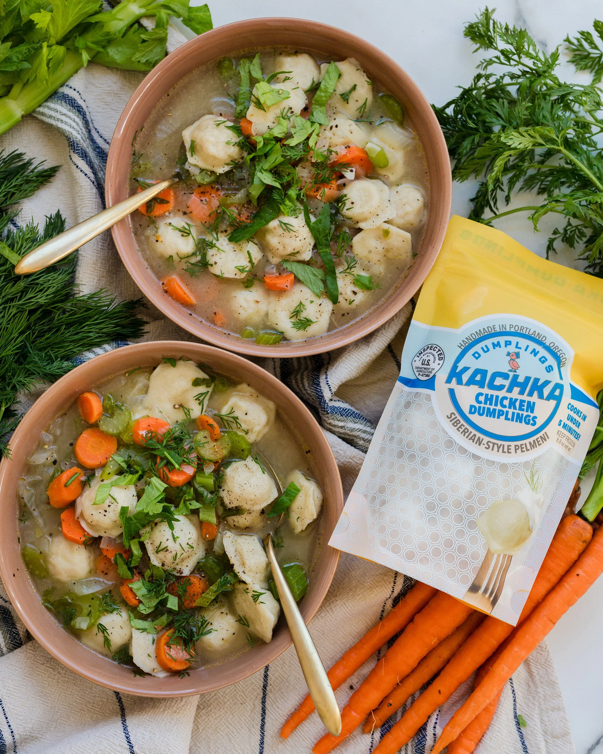 Two bowls of chicken dumpling soup garnished with fresh herbs, surrounded by fresh carrots, herbs, and a package of Kachka chicken dumplings.